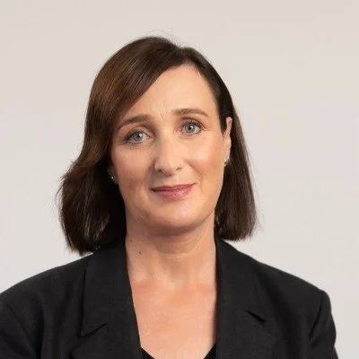 Portrait of a woman with shoulder-length brown hair wearing a black blazer, smiling against a plain light background.