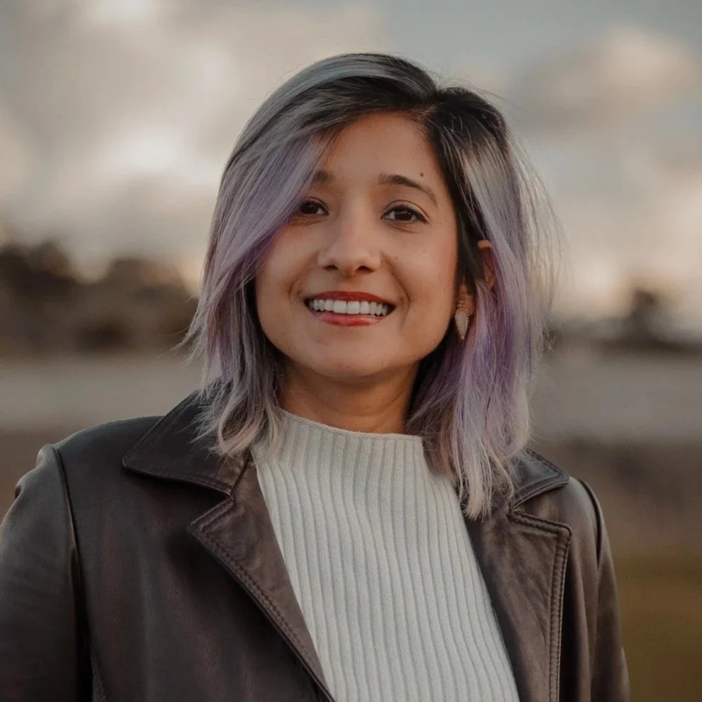 A woman with shoulder-length black hair, wearing a green jacket with purple floral designs over a black top, smiling indoors against a beige wall.