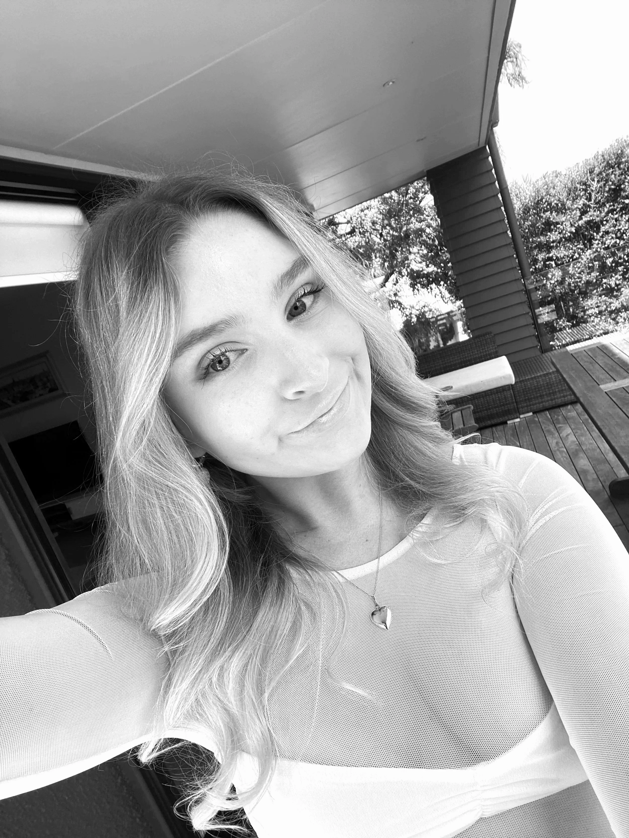 Black and white photo of a young woman with long wavy hair, smiling, wearing a light-colored top and a necklace with a heart pendant, standing outdoors near a patio with a wooden deck and trees in the background.