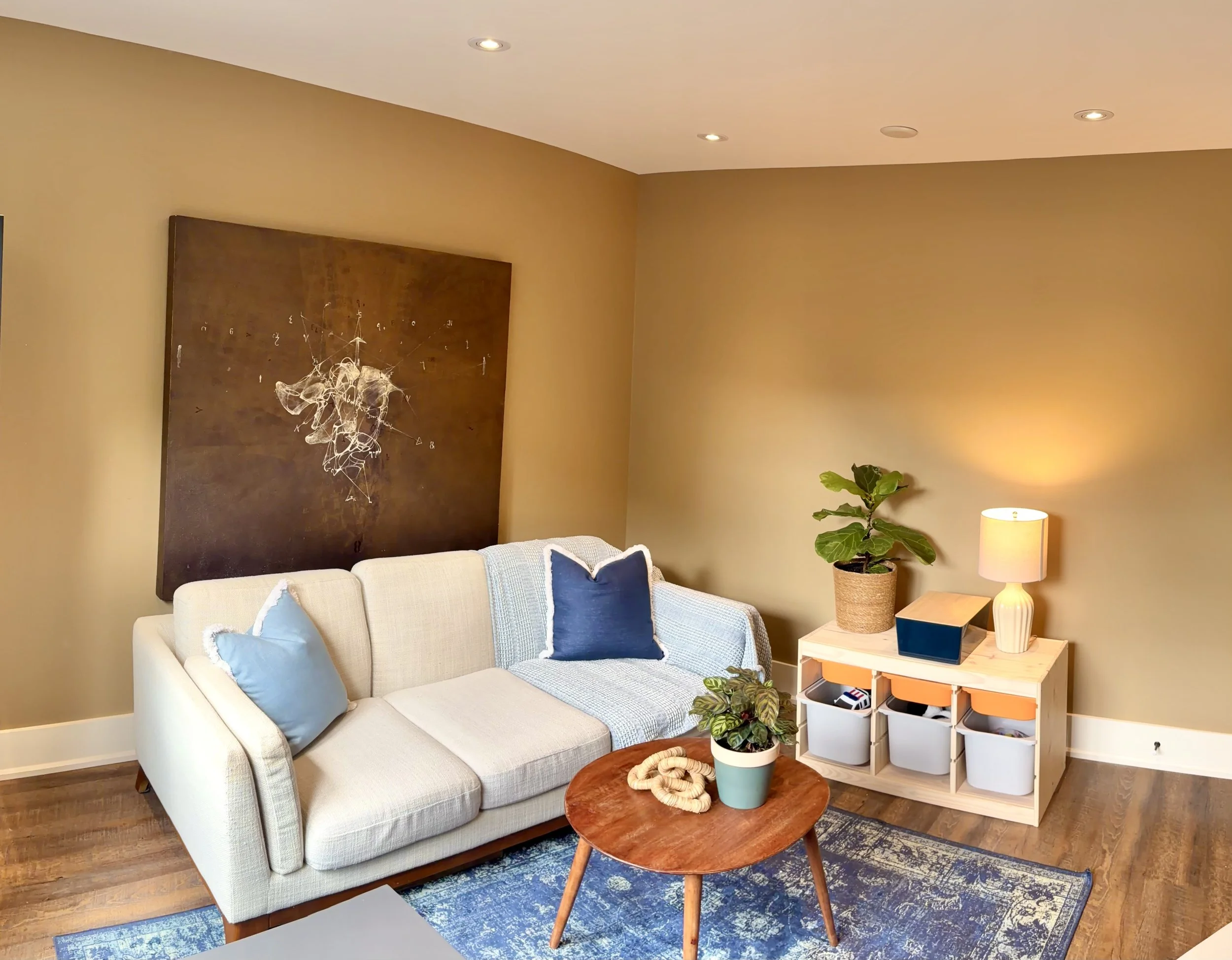 Living room with beige couch, blue and beige throw pillows, large abstract artwork on the wall, plant in a pot, white side table with lamp, and decorative items on the table, hardwood floor with blue patterned area rug.