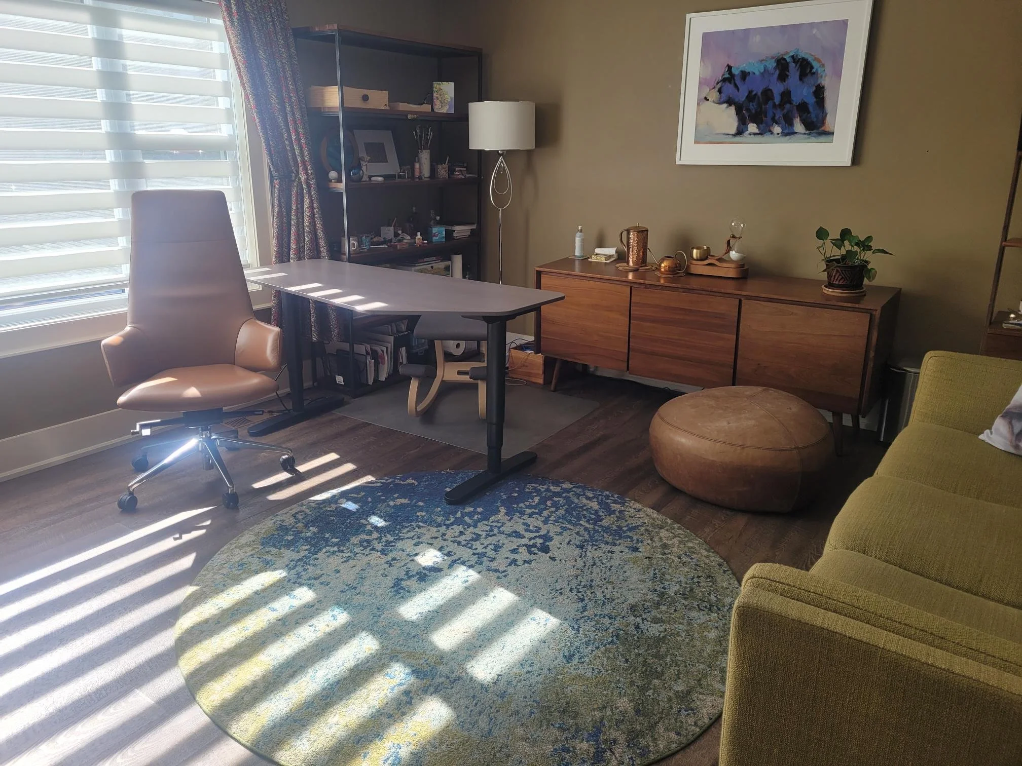 A cozy living room with a window on the left, a beige swivel chair, a round blue and yellow patterned rug, a leather pouf, a wooden sideboard with decorative items, and a framed painting of a bear on the wall.