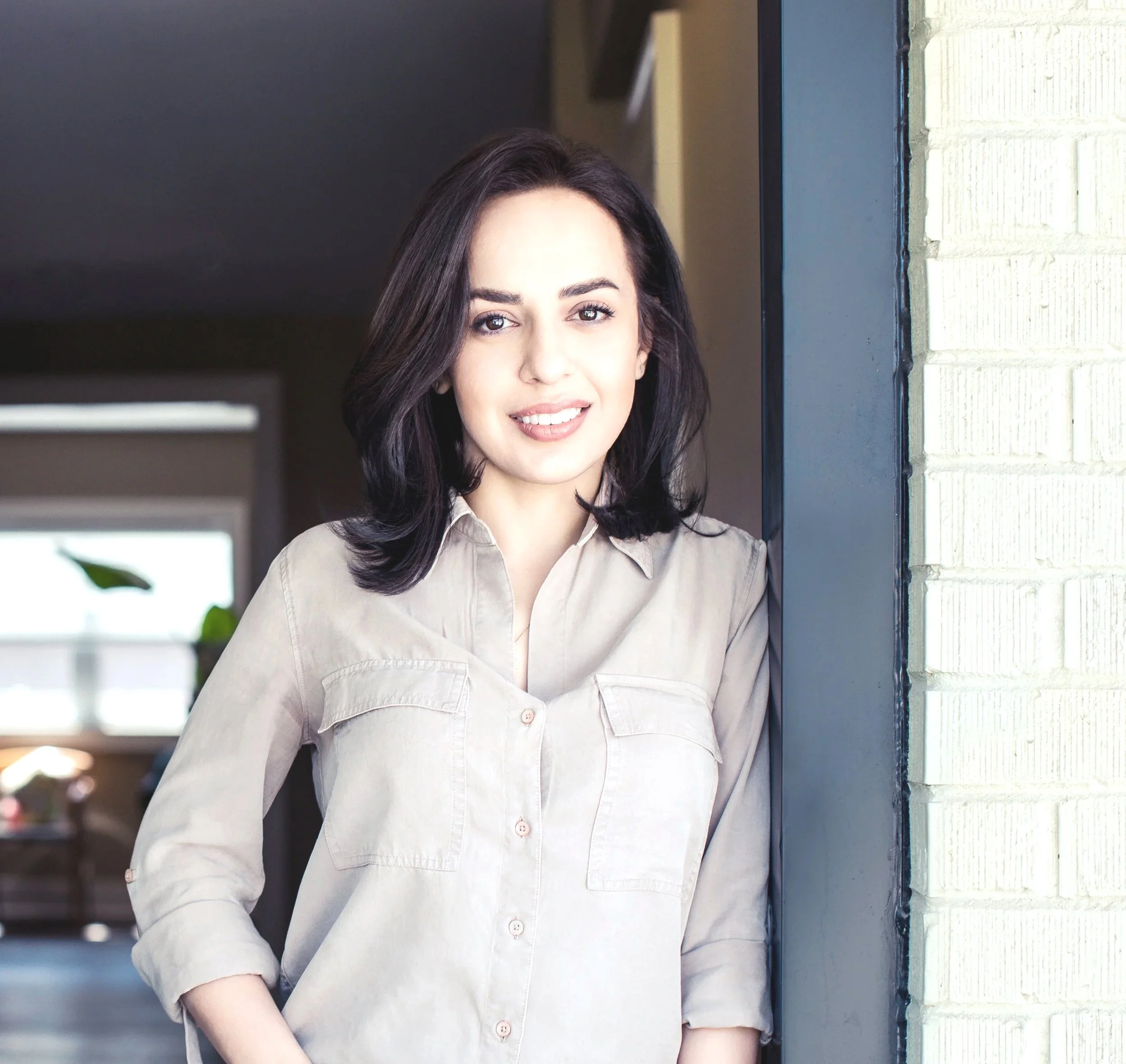 A woman with shoulder-length dark hair and light skin smiling, wearing a beige button-up shirt, standing outdoors next to a brick wall.