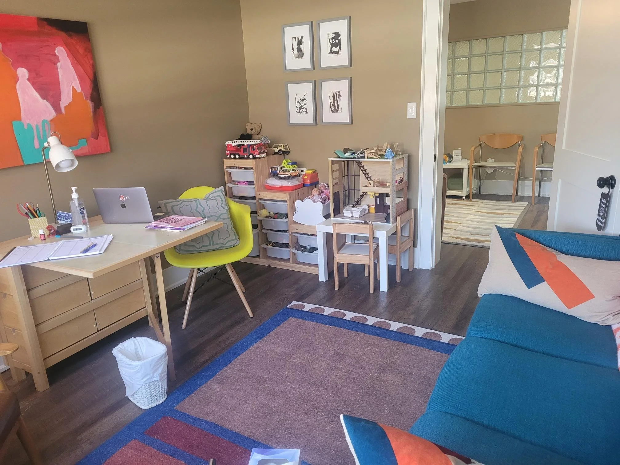 A spacious room with a beige wall featuring four framed black and white abstract artworks. There is a family room with a green chair, a white table holding a laptop, notebooks, and a lamp, and a small wooden dollhouse with toys nearby. An open doorway leads to another room with two chairs and a window wall. The room has a multi-colored area rug and various children's toys and art supplies.