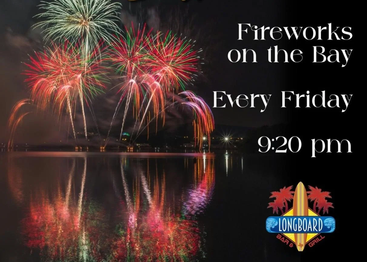 Fireworks display over a bay reflecting colorful lights on the water, with promotional text for weekly fireworks shows on Fridays at 9:20 pm at Longboard Bar & Grill, featuring a logo with a surfboard and palm trees on south padre island