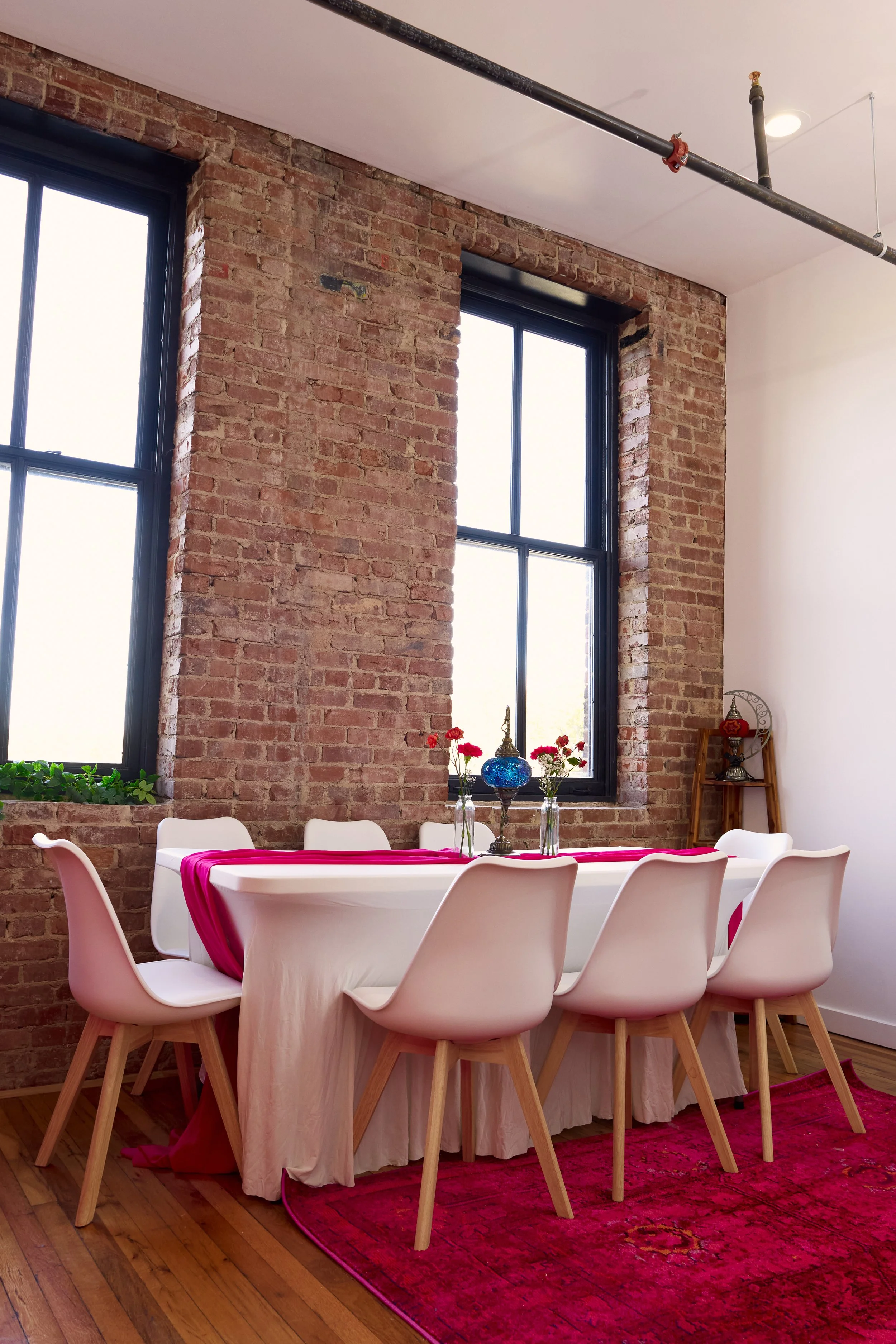 A dining area with a white table, six white chairs with wooden legs, a pink table runner, and three glass vases with red flowers. The room has exposed brick walls, large windows, and a red rug on wooden floor.