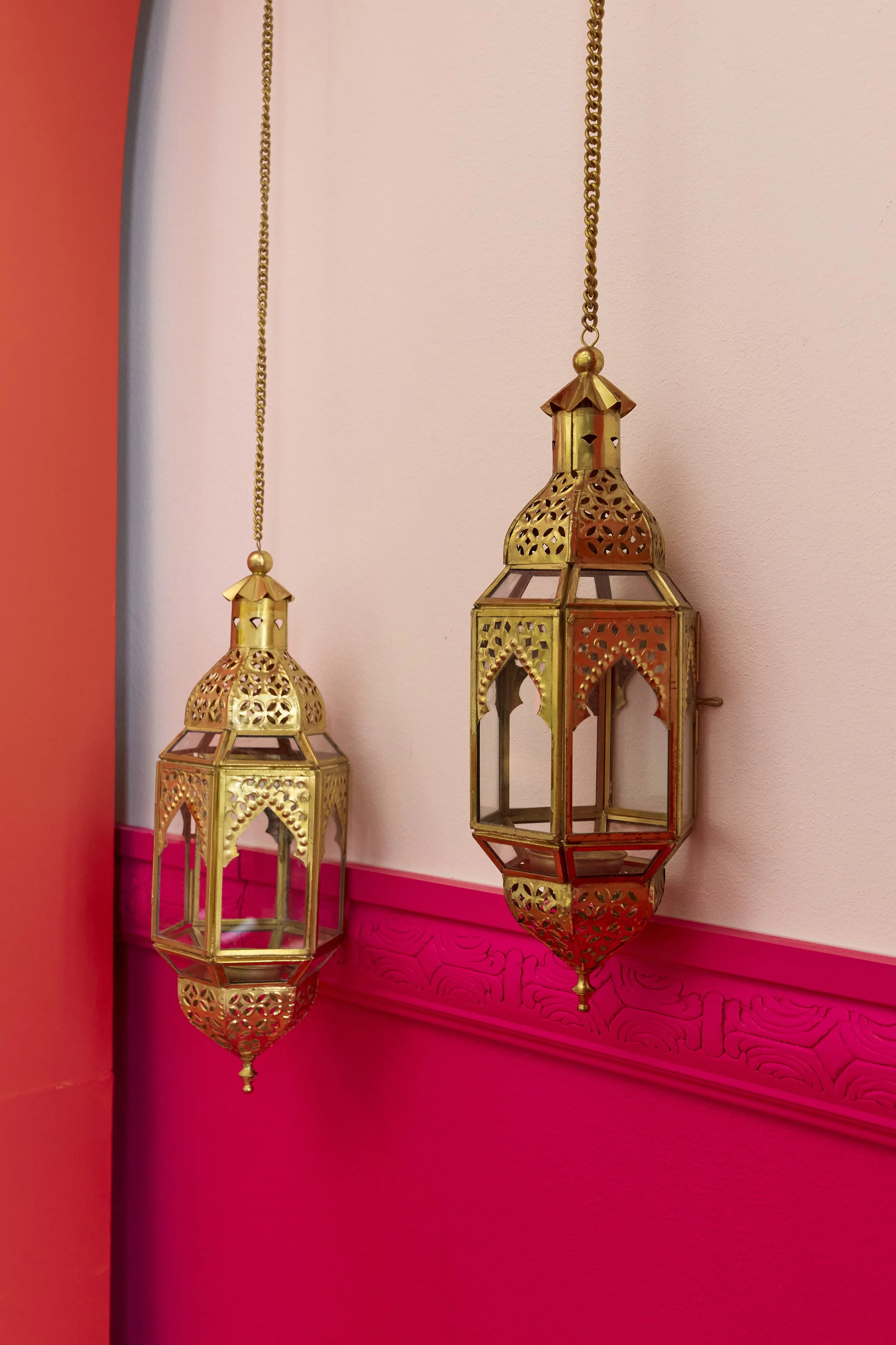 Two ornate brass lanterns hanging from gold chains on a pink wall with pink decorative molding.