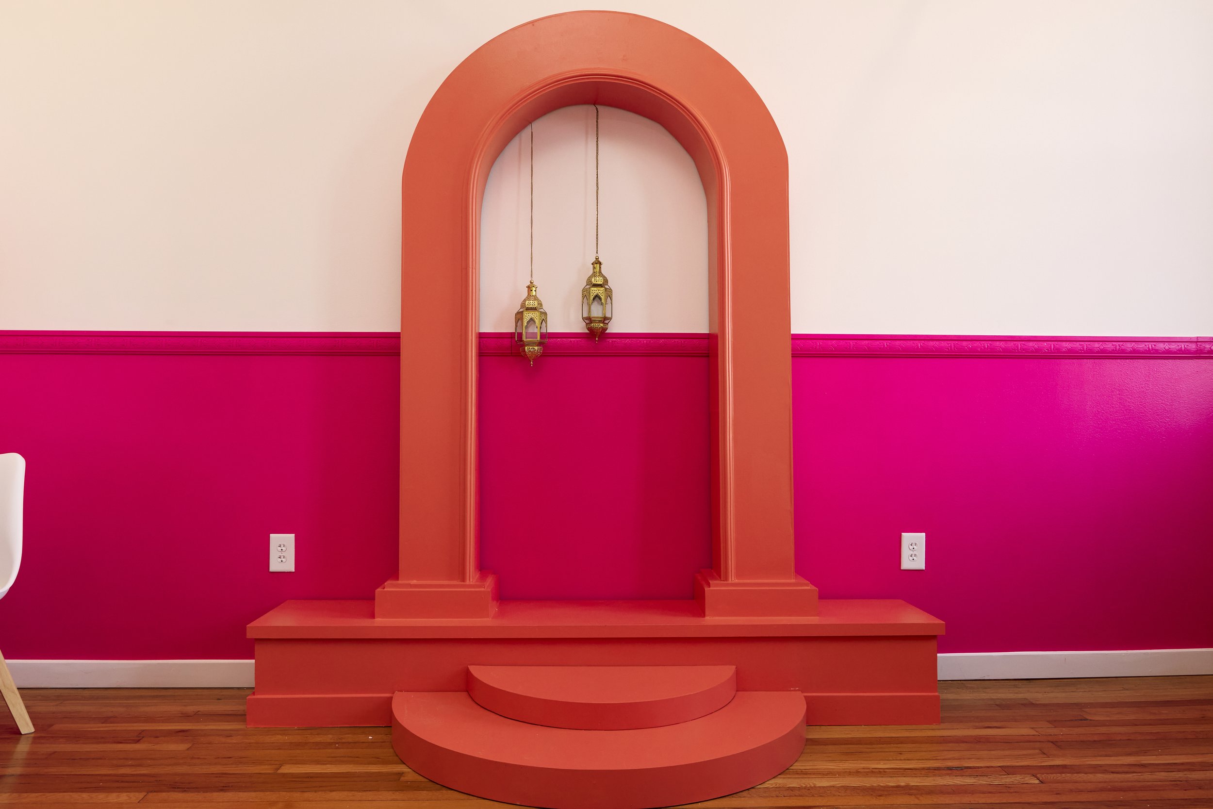 Decorative pink wall with coral arch, two hanging lanterns, and a small set of steps at the bottom, wooden floor.