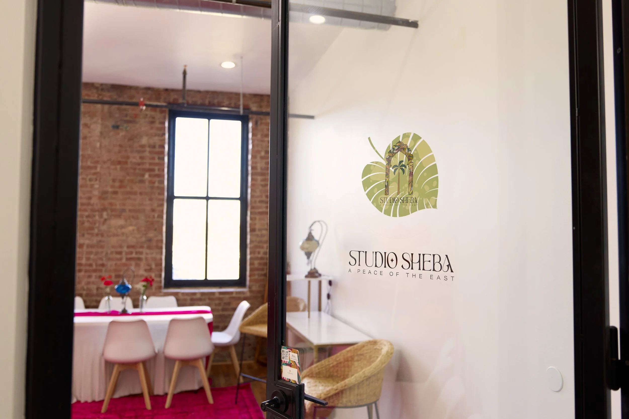 The image shows the entrance to Studio Sheba, with a glass door displaying the logo and name. Inside, there is a dining area with a table covered with a pink cloth, surrounded by white and pink chairs, and decorated with small vases and flowers. The 