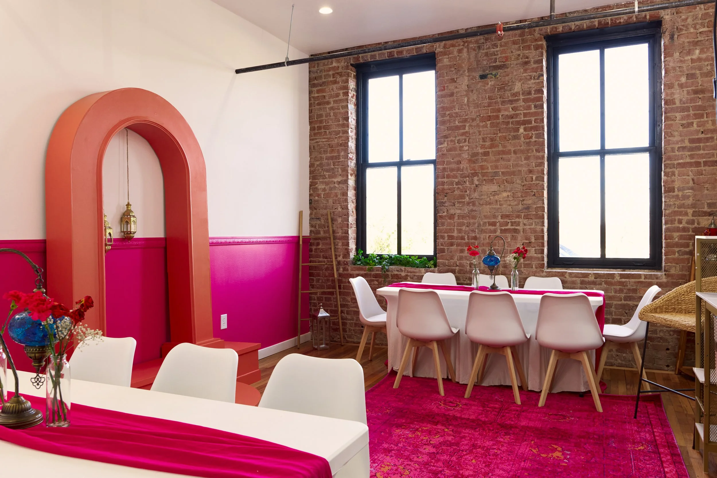 Interior of a decorated dining room with pink and white accents, exposed brick walls, large black-framed windows, white chairs around tables with pink tablecloths, and pink carpeting.