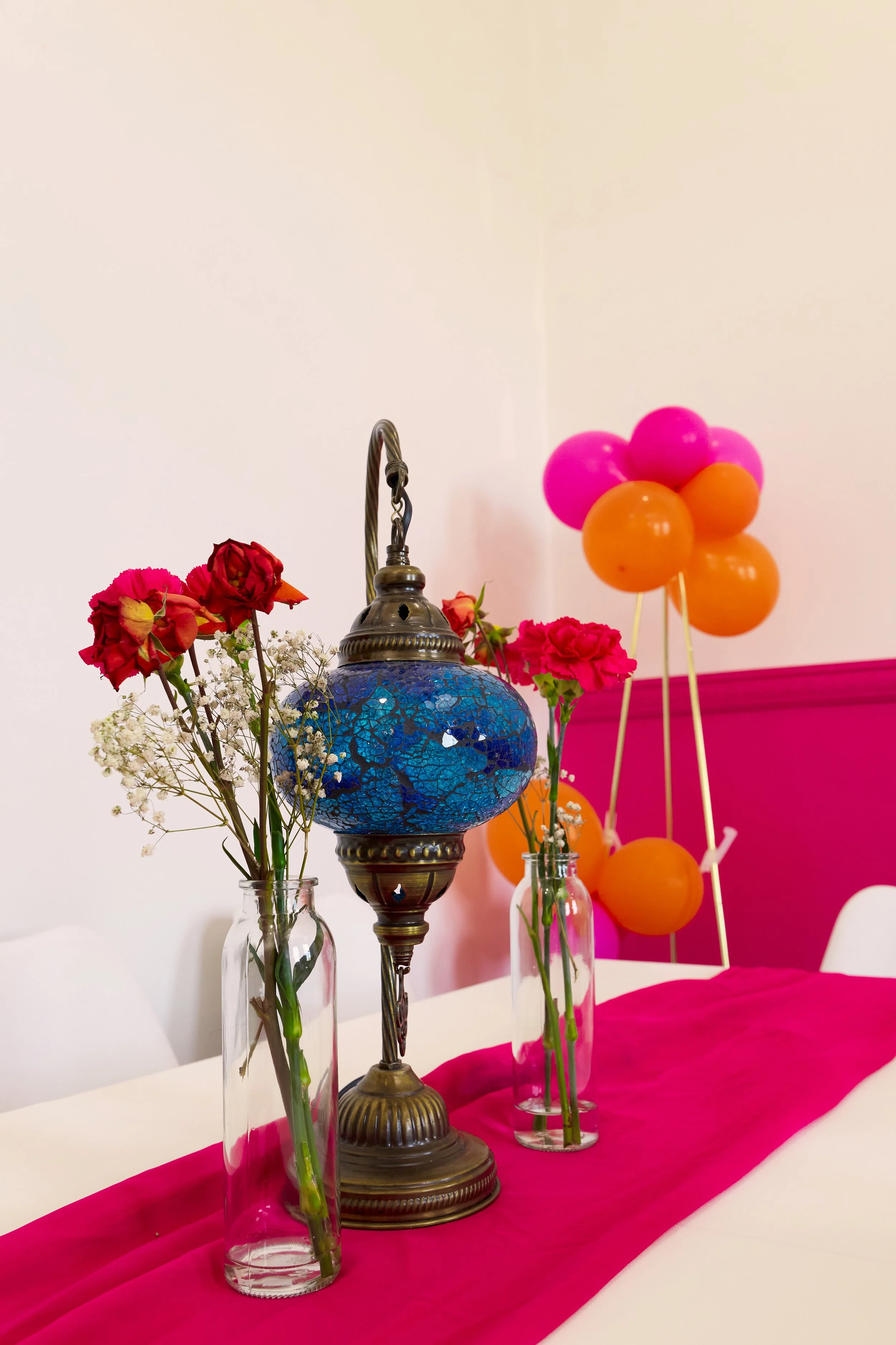 Decorative table setting with pink and orange balloons, pink flowers, white baby's breath, and a vintage lamp with blue glass mosaic, on a pink table runner