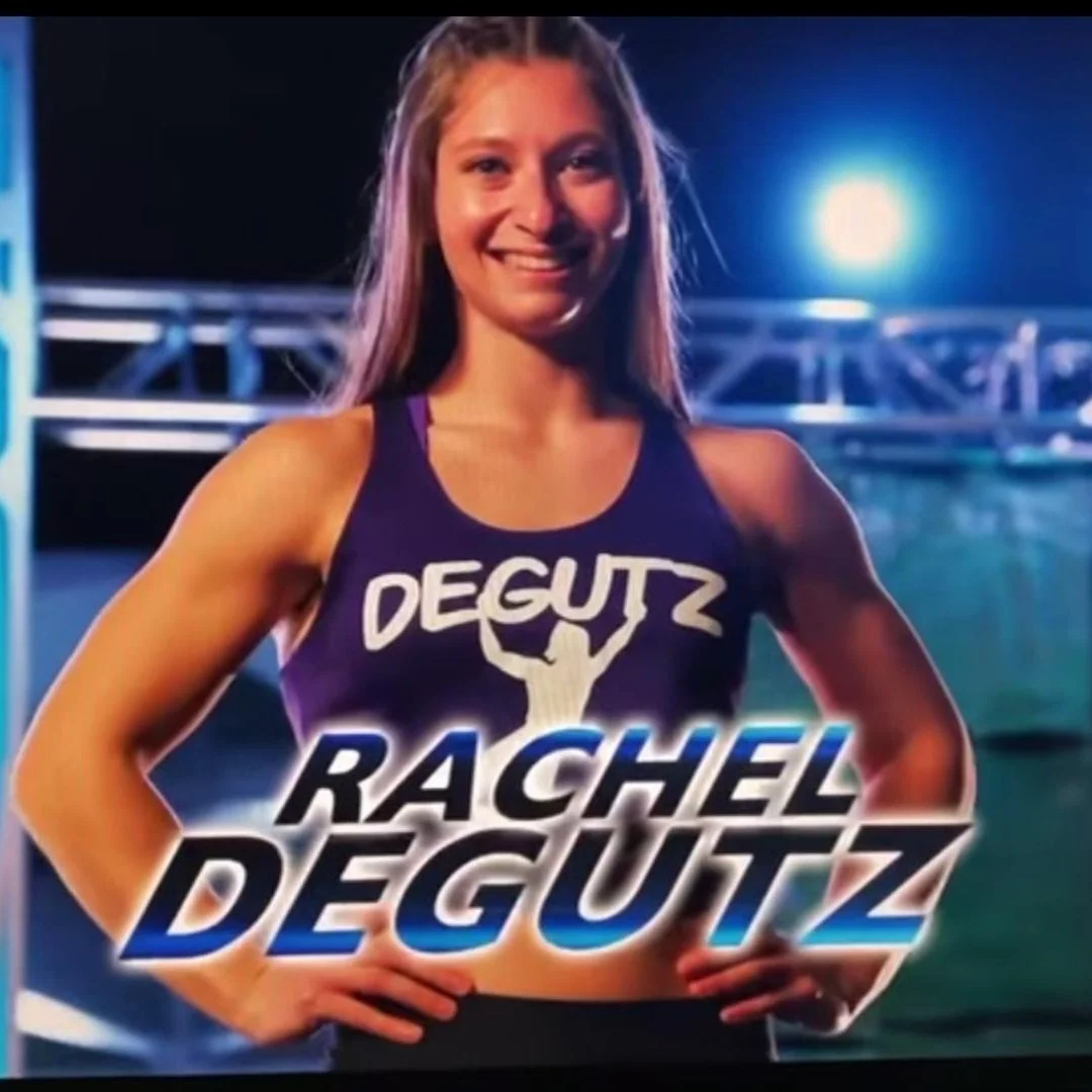 A young woman in a purple athletic sports bra stands with her hands on her hips. She has long hair and is smiling. The background appears to be a nighttime outdoor setting with bright lights and a metal structure. Text on the image reads "DEGUTZ" and