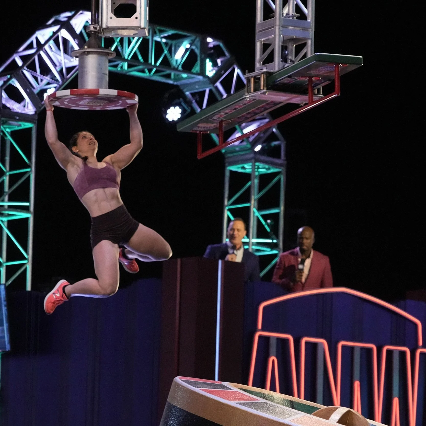 A female athlete in athletic wear is mid-air, holding a round object, possibly a disc, during a game or competition in an indoor arena with bright neon lights and a metal structure overhead. Two men with microphones watch from behind a dark blue barrier.
