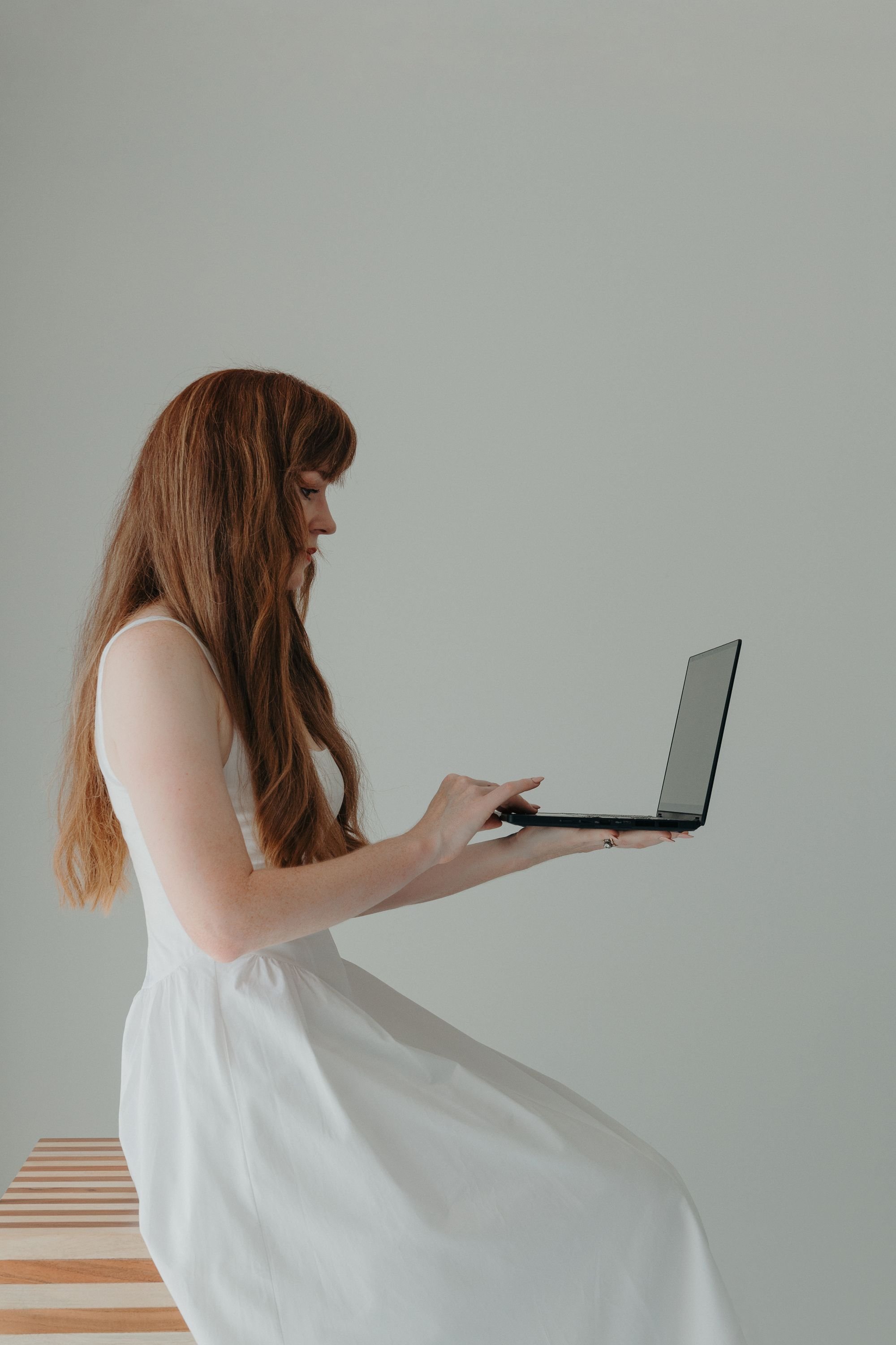 A young woman with long red hair sitting on a striped wooden bench, wearing a white dress and using a laptop against a plain light green background.