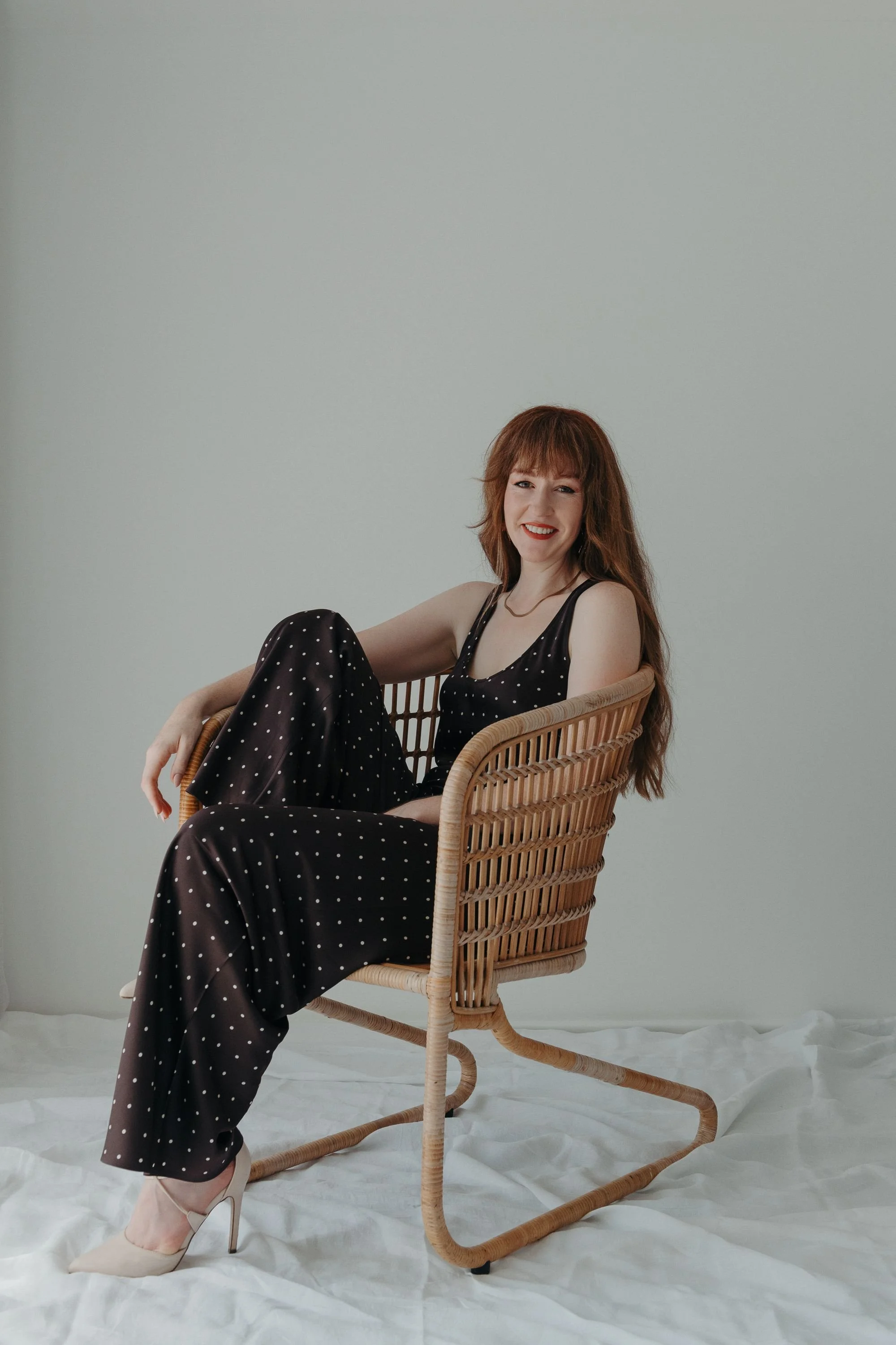 A young woman with long red hair and a smile, wearing a black polka dot jumpsuit, sitting on a wicker chair with a white backdrop.