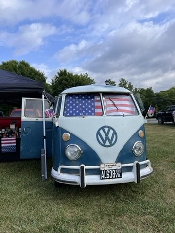 A vintage blue Volkswagen van with American flags displayed in the windows and on poles. It's parked on grass beside a black canopy with a partially open side door showing a glimpse of supplies. The weather is partly cloudy.