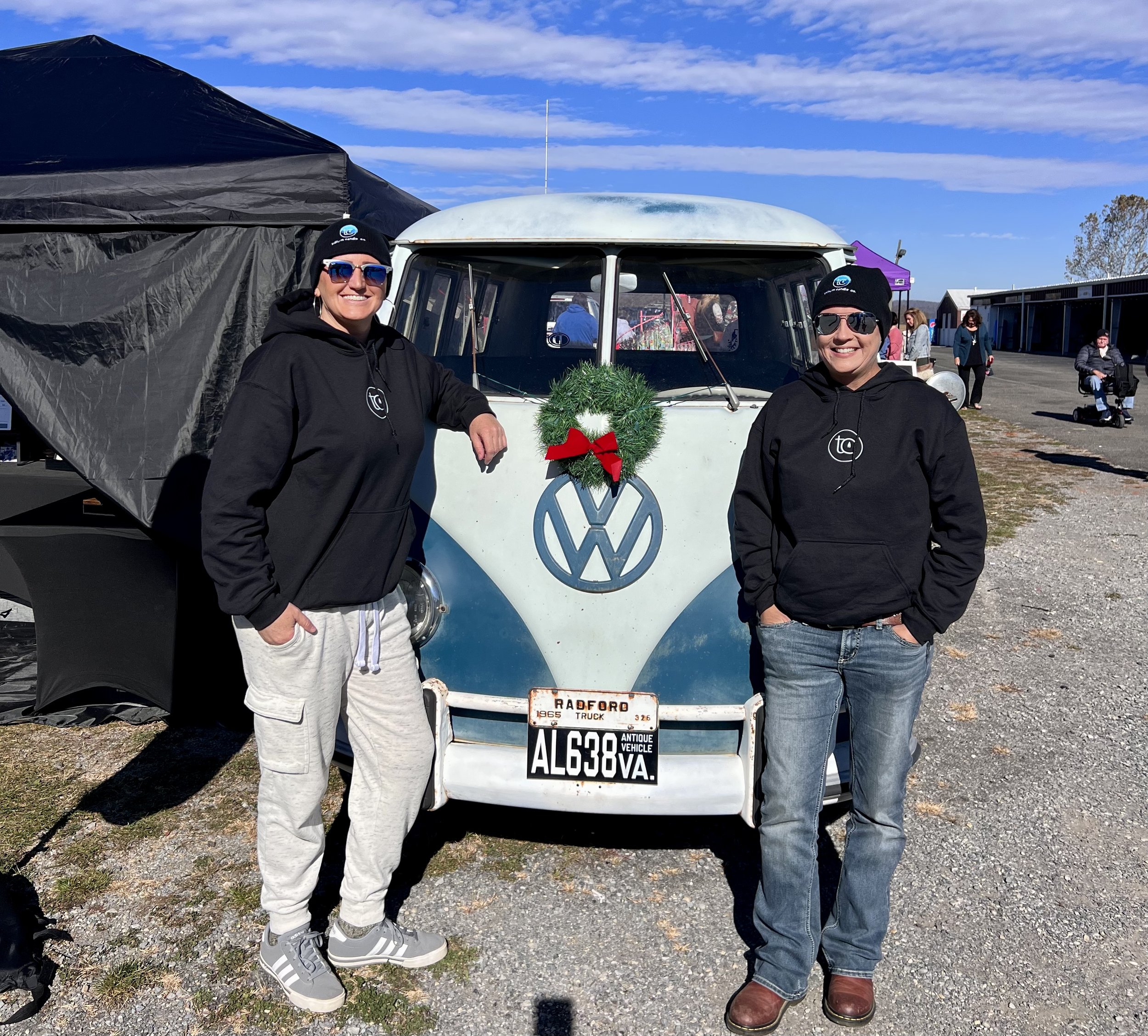 Two people wearing black hoodies and hats standing in front of a vintage blue and white Volkswagen van with a wreath on the front. The van has a Virginia license plate and is parked on a gravel surface. A canopy is set up nearby under a blue sky.