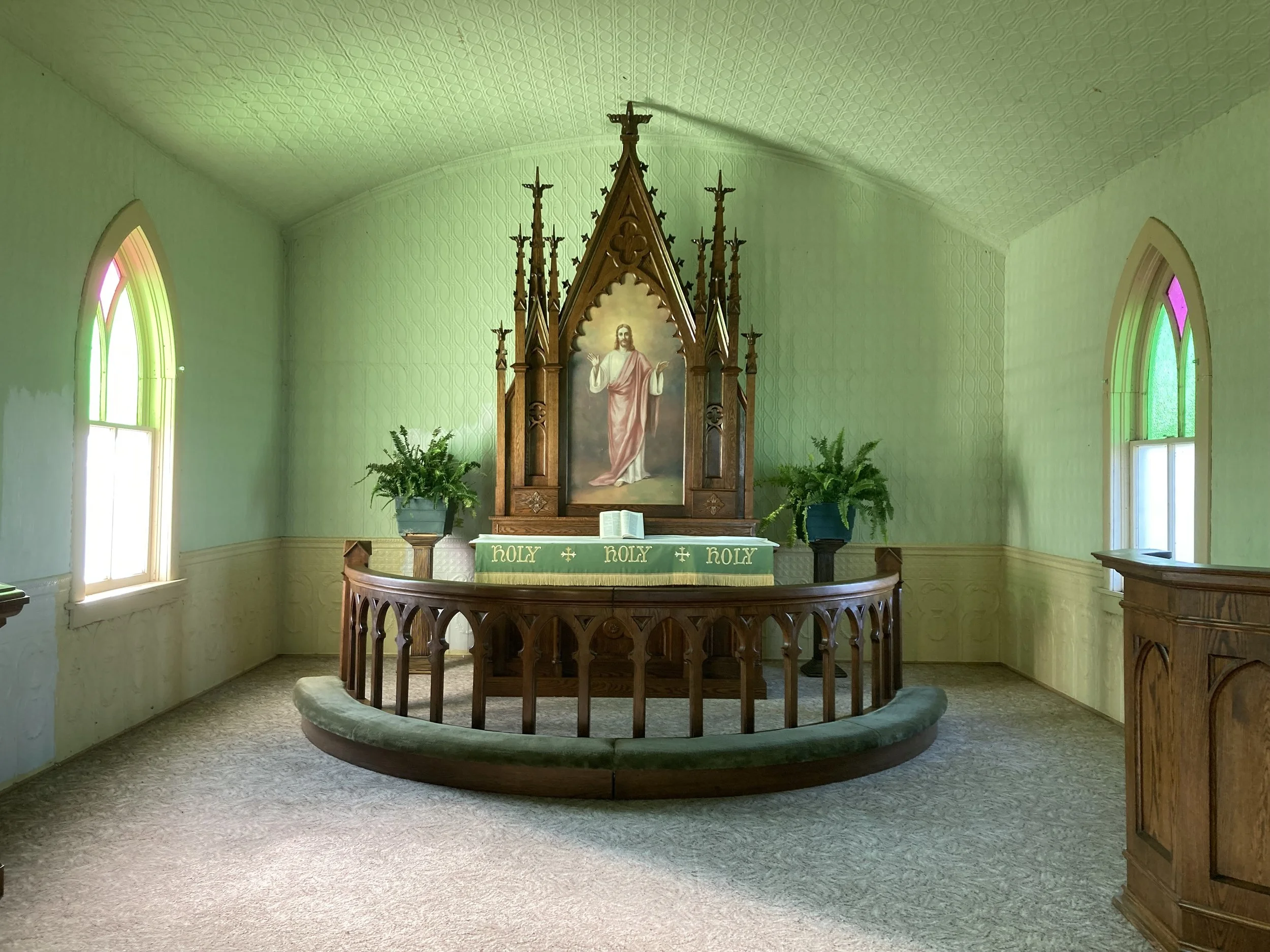Altar space of Tunbridge Lutheran Church with stained glass windows, and an altar featuring a painting of Jesus Christ with open arms draped with green seasonal vestments.