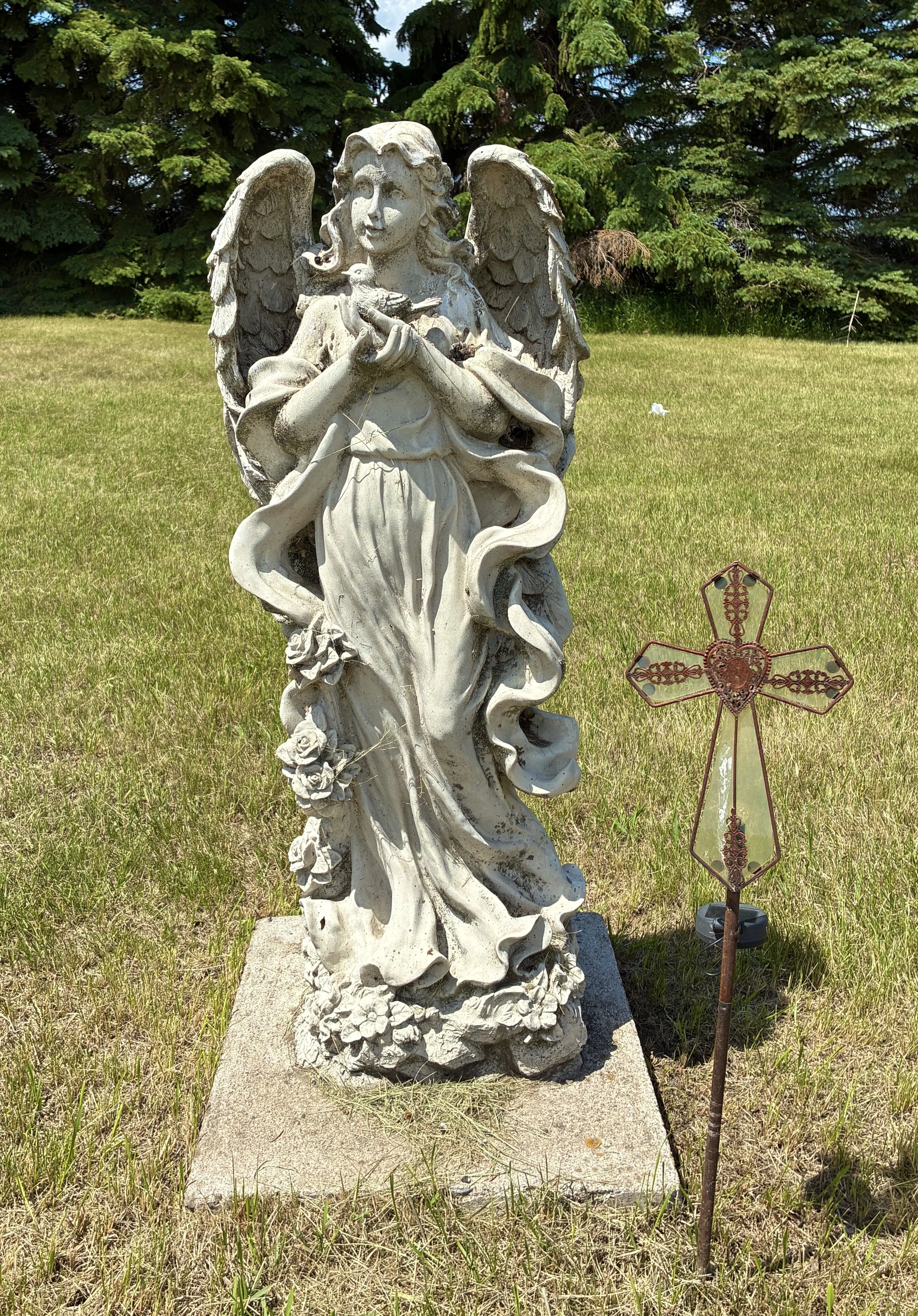 A stone angel statue with wings, holding a dove, standing on a stone pedestal in Tunbridge Lutheran Cemetery with tall trees in the background, and a decorative metal cross nearby.