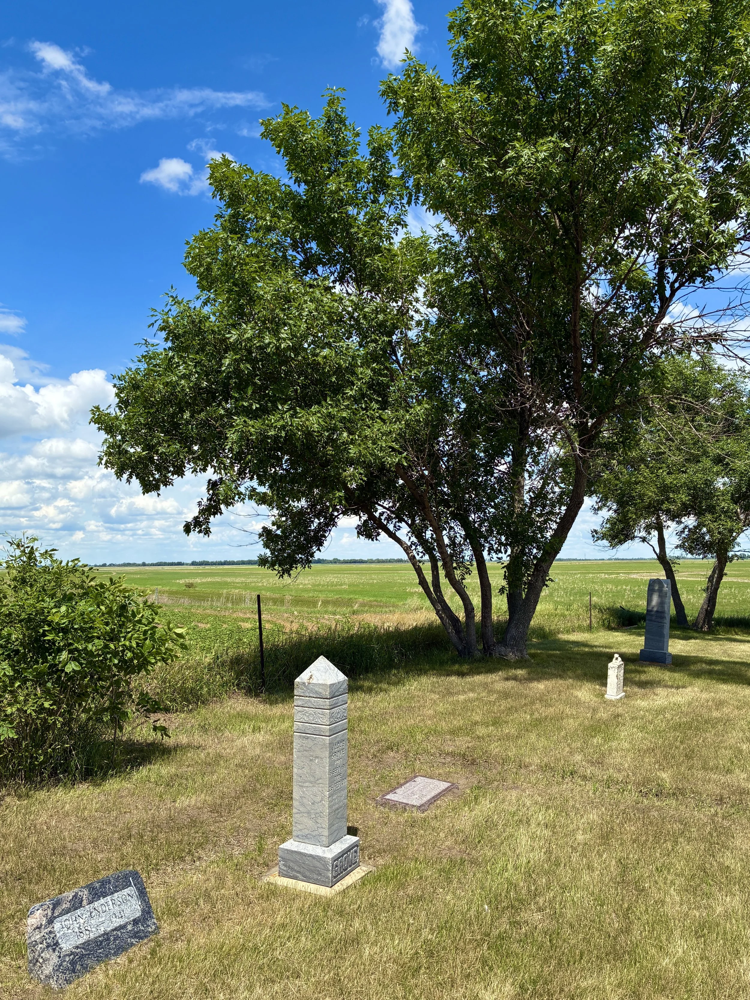 View of the northwest corner of Tunbridge Lutheran Church with several tombstones in the grass, a large green-leafed tree, and a bright blue sky with scattered clouds.