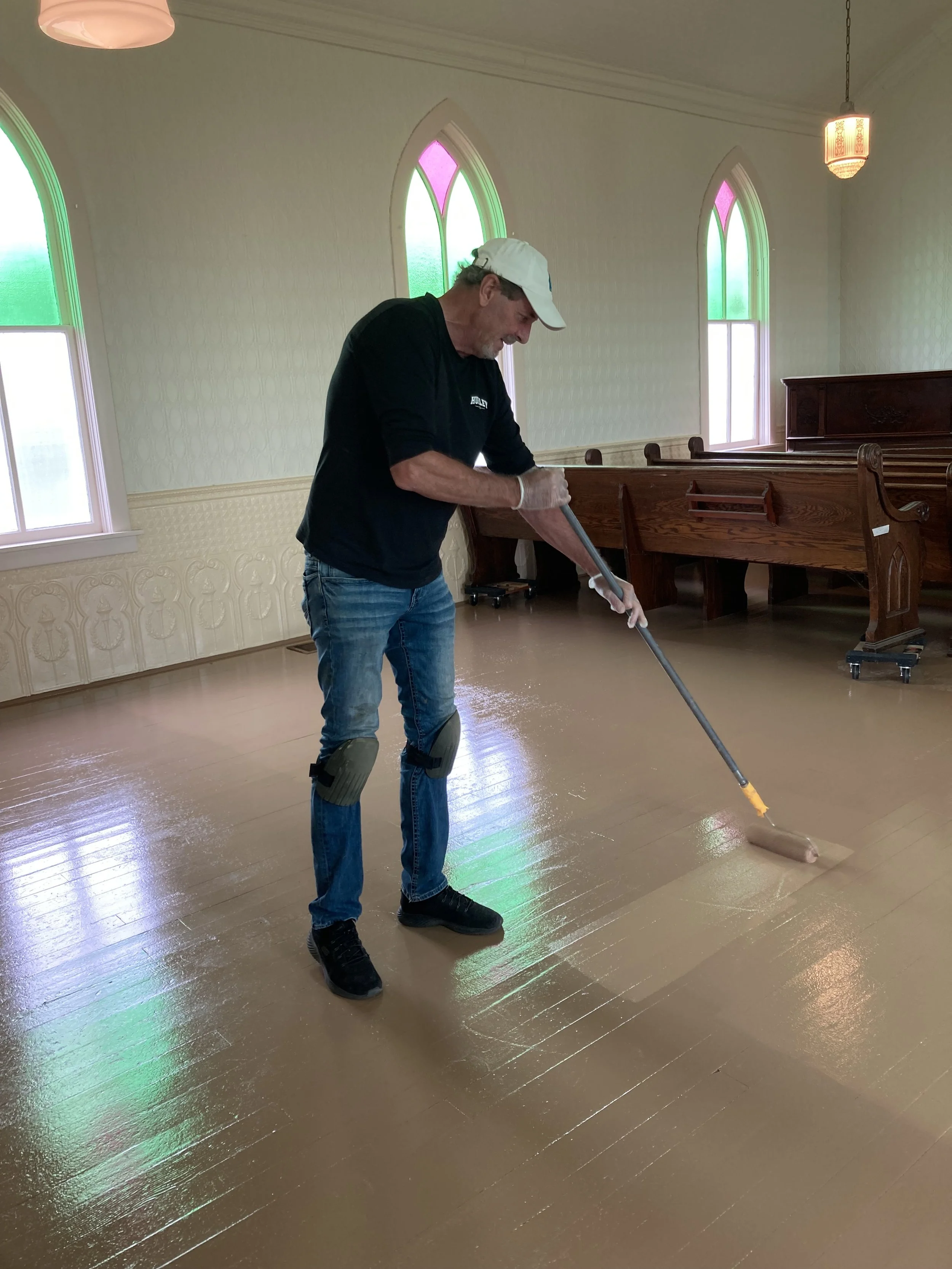 A man wearing a white cap, black sweatshirt, jeans, and black shoes uses a roller to paint the floor inside the Tunbridge Lutheran Church sanctuary. 