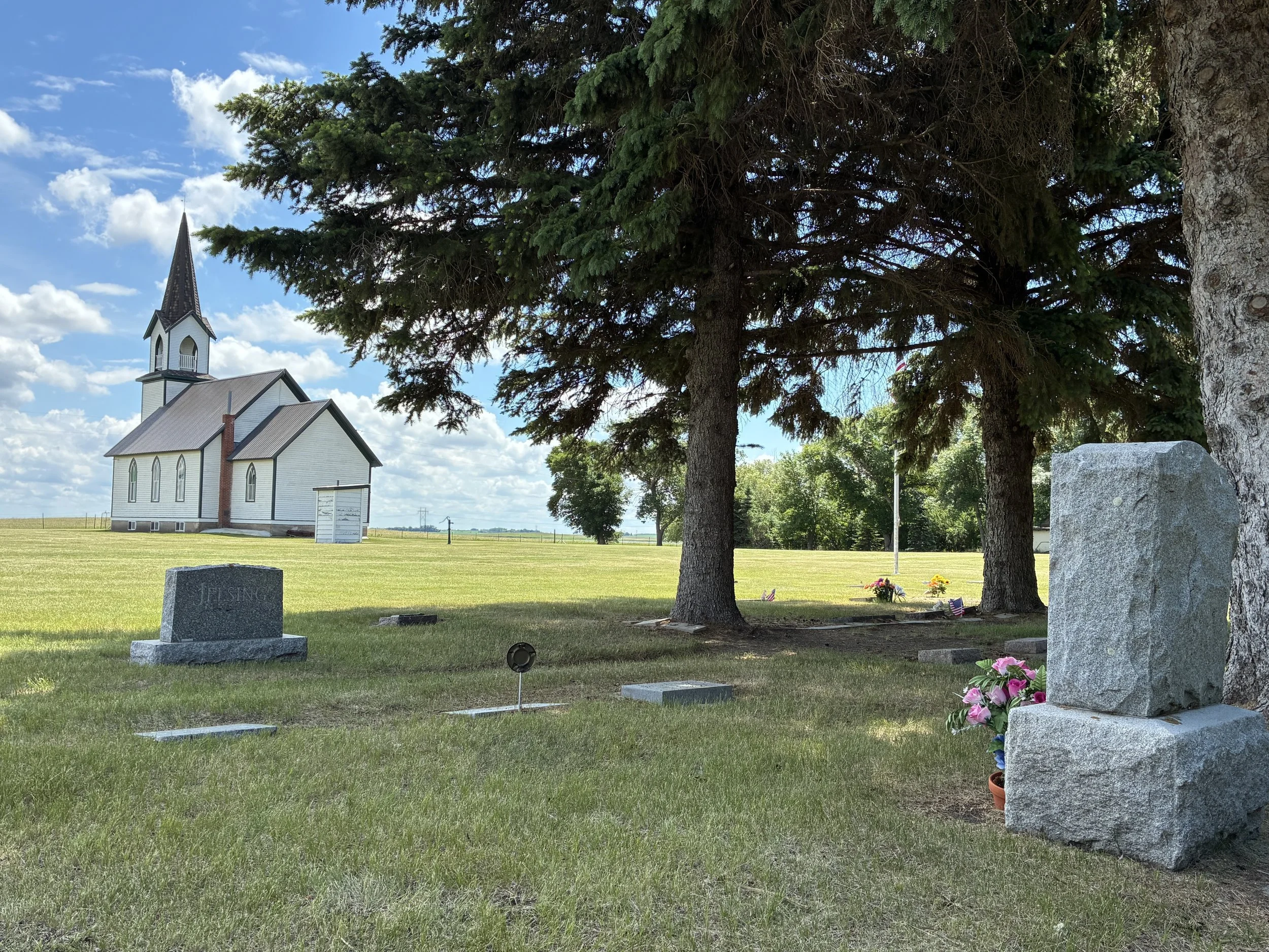 View of Tunbridge Lutheran Church with its white siding and tall steeple, surrounded by a grassy cemetery with headstones, some with flowers, under a partly cloudy sky, with large trees in the foreground.