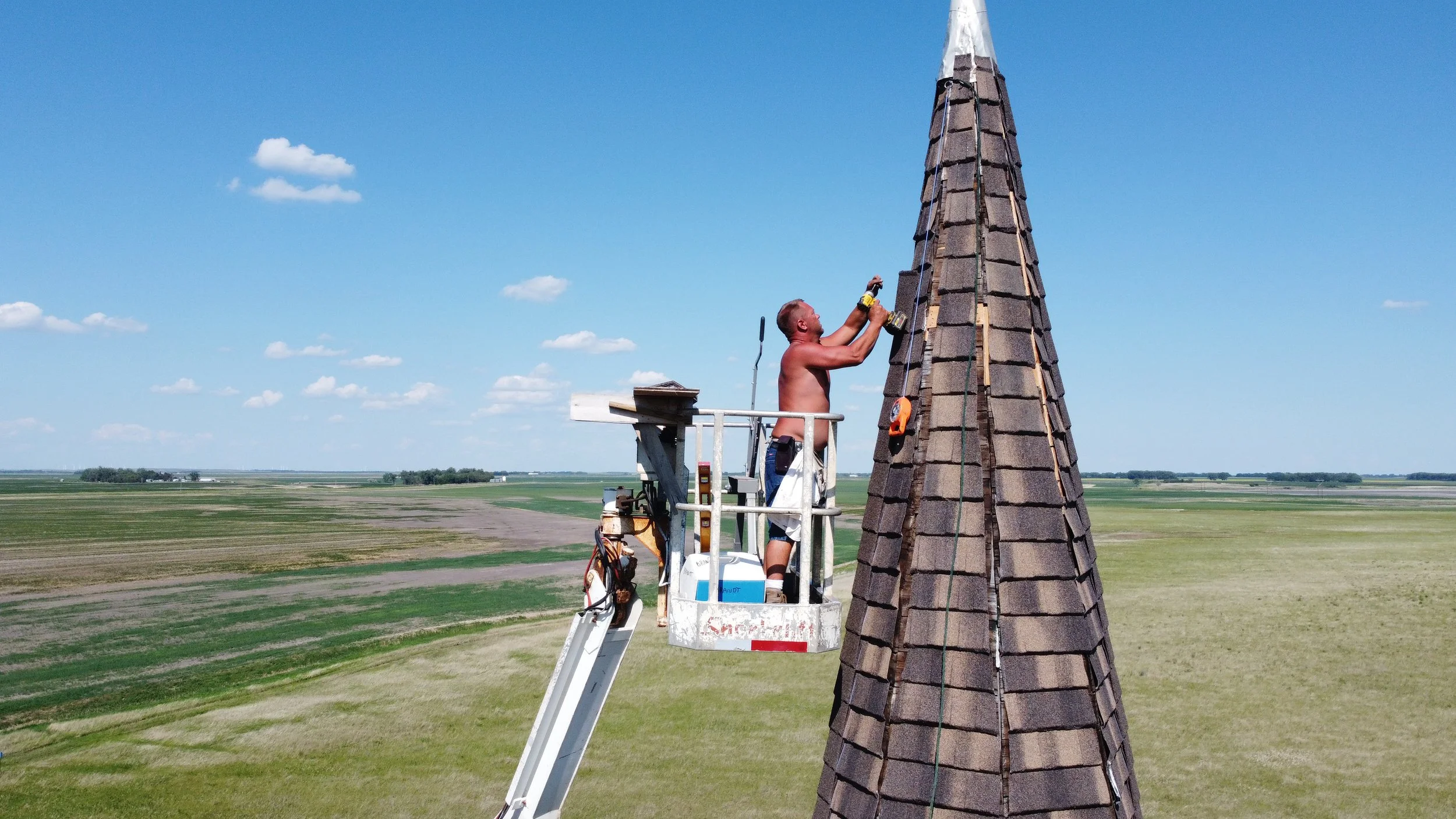A man uses a lift to shingle the Tunbridge Lutheran Church steeple, under a blue sky with some clouds, with open farmland in the background.
