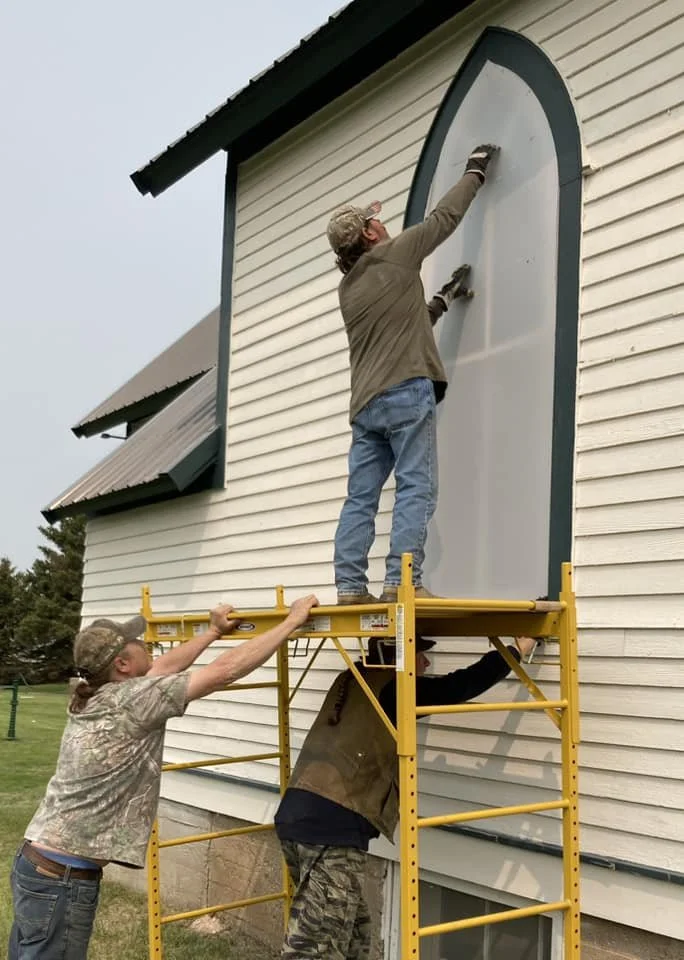 A volunteer covers a stained glass window at Tunbridge Lutheran Church with a protective shield, while two volunteers support the scaffolding from below.