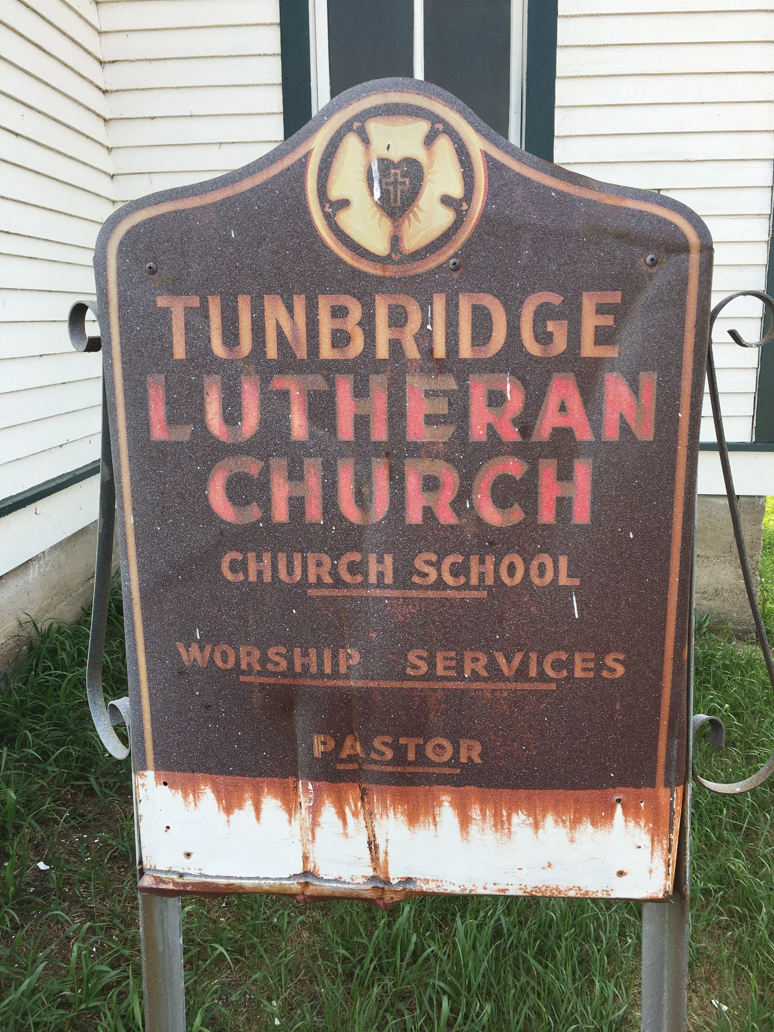 A rusted, weathered sign for Tunbridge Lutheran Church and Church School, featuring a heart with a cross with information on worship services .