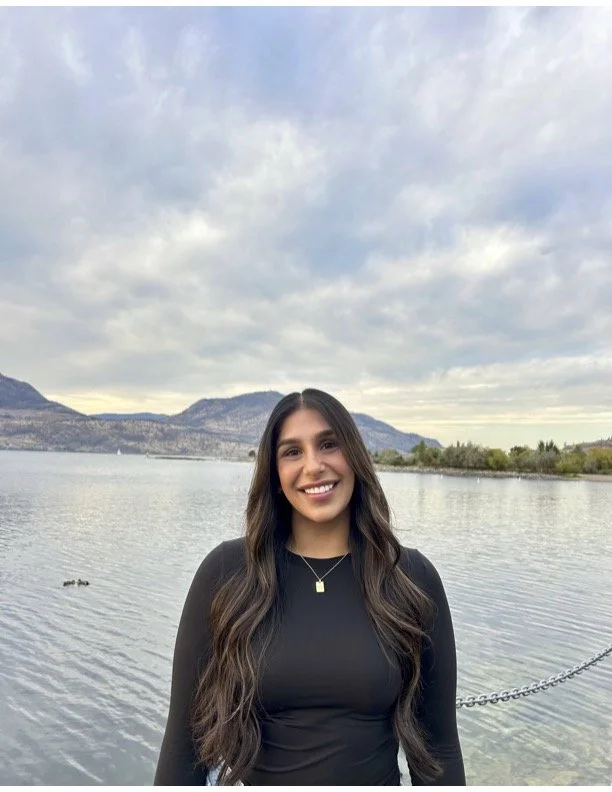 A young woman with long brown hair smiles outdoors near a lake, with mountains and a cloudy sky in the background.