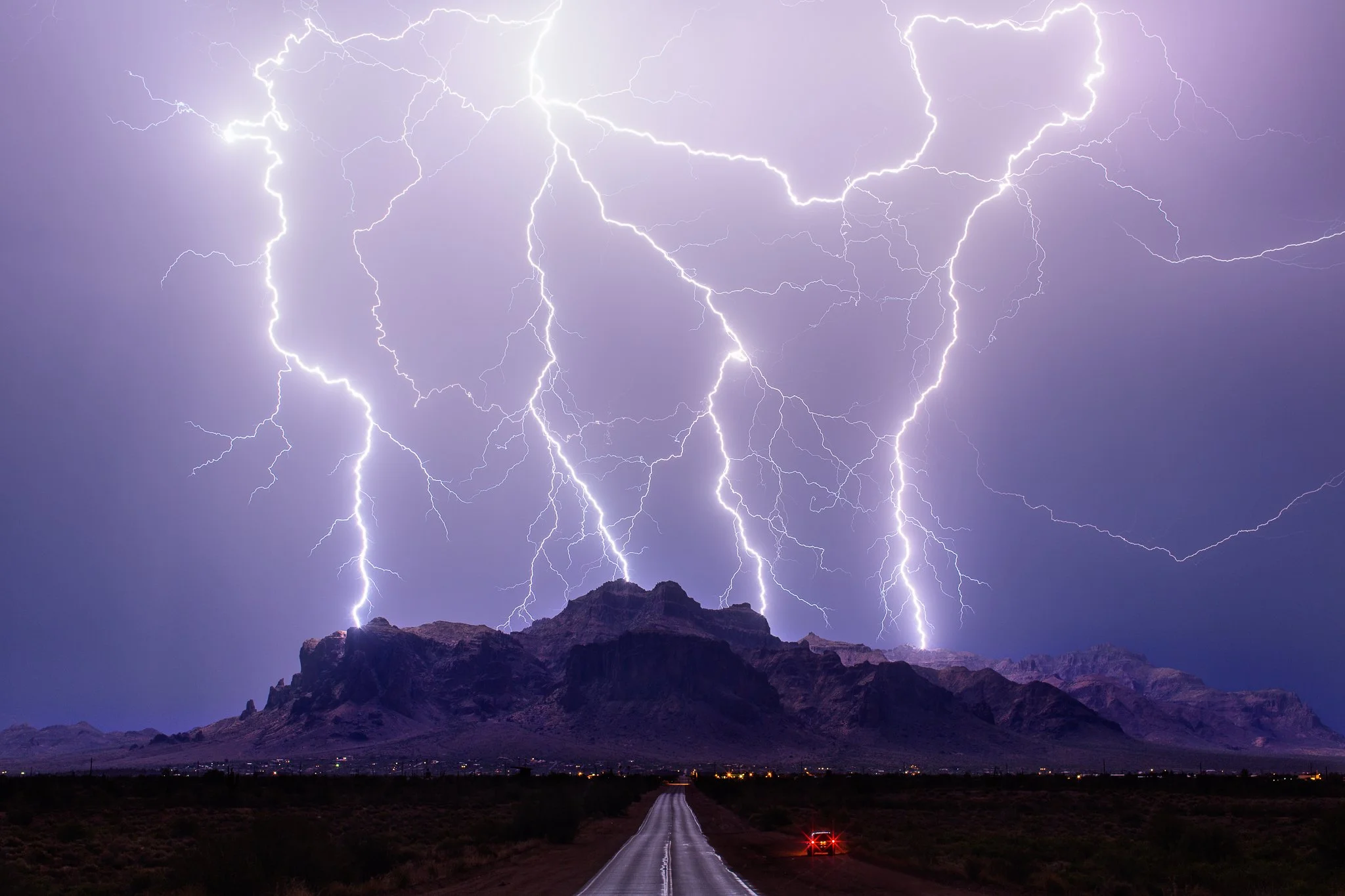 Dramatic thunderstorm over a mountain with multiple lightning strikes illuminating the night sky, a road leading towards the mountains, and a car's taillights visible.