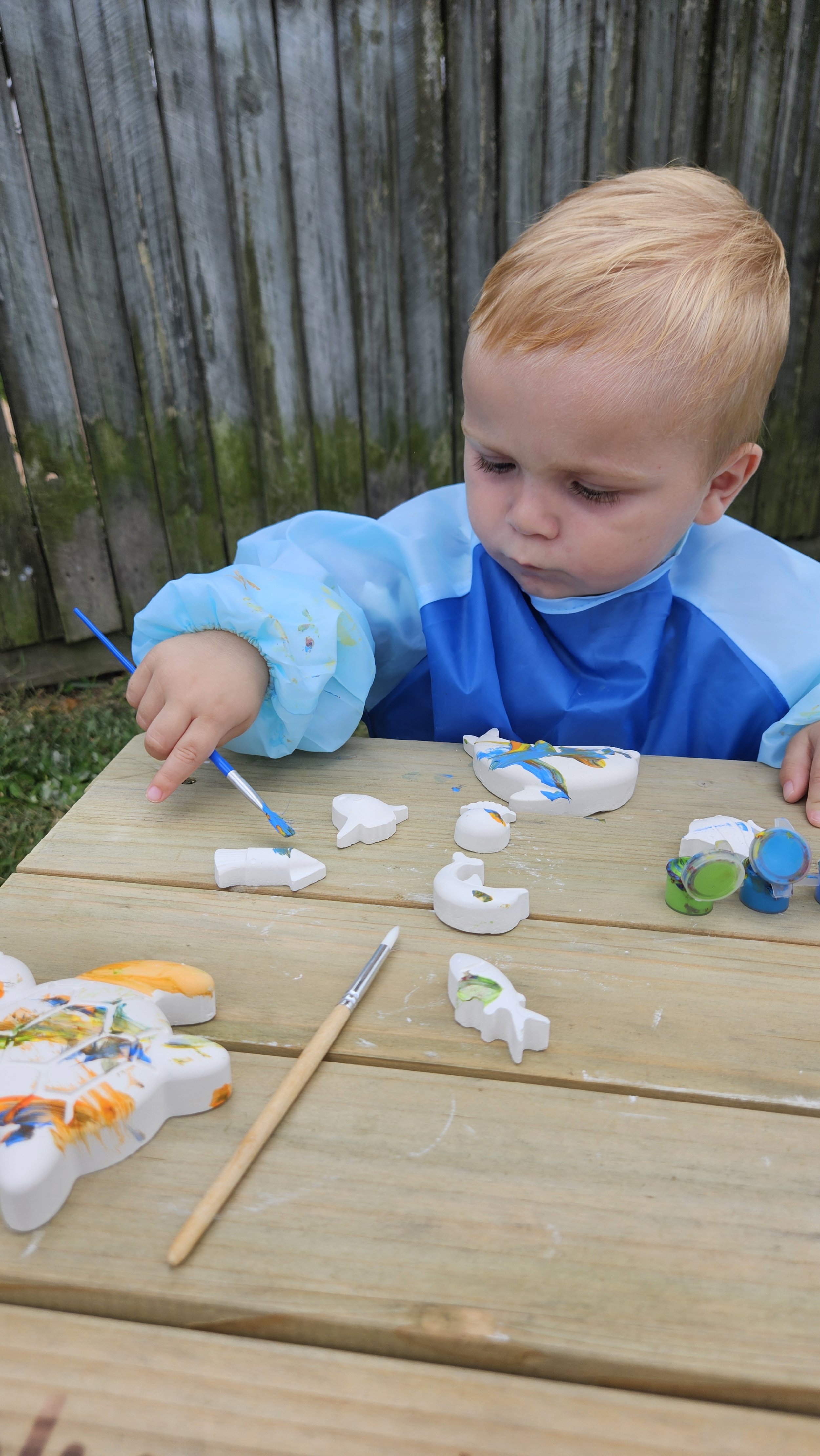Child painting plaster shapes on a wooden table outdoors.