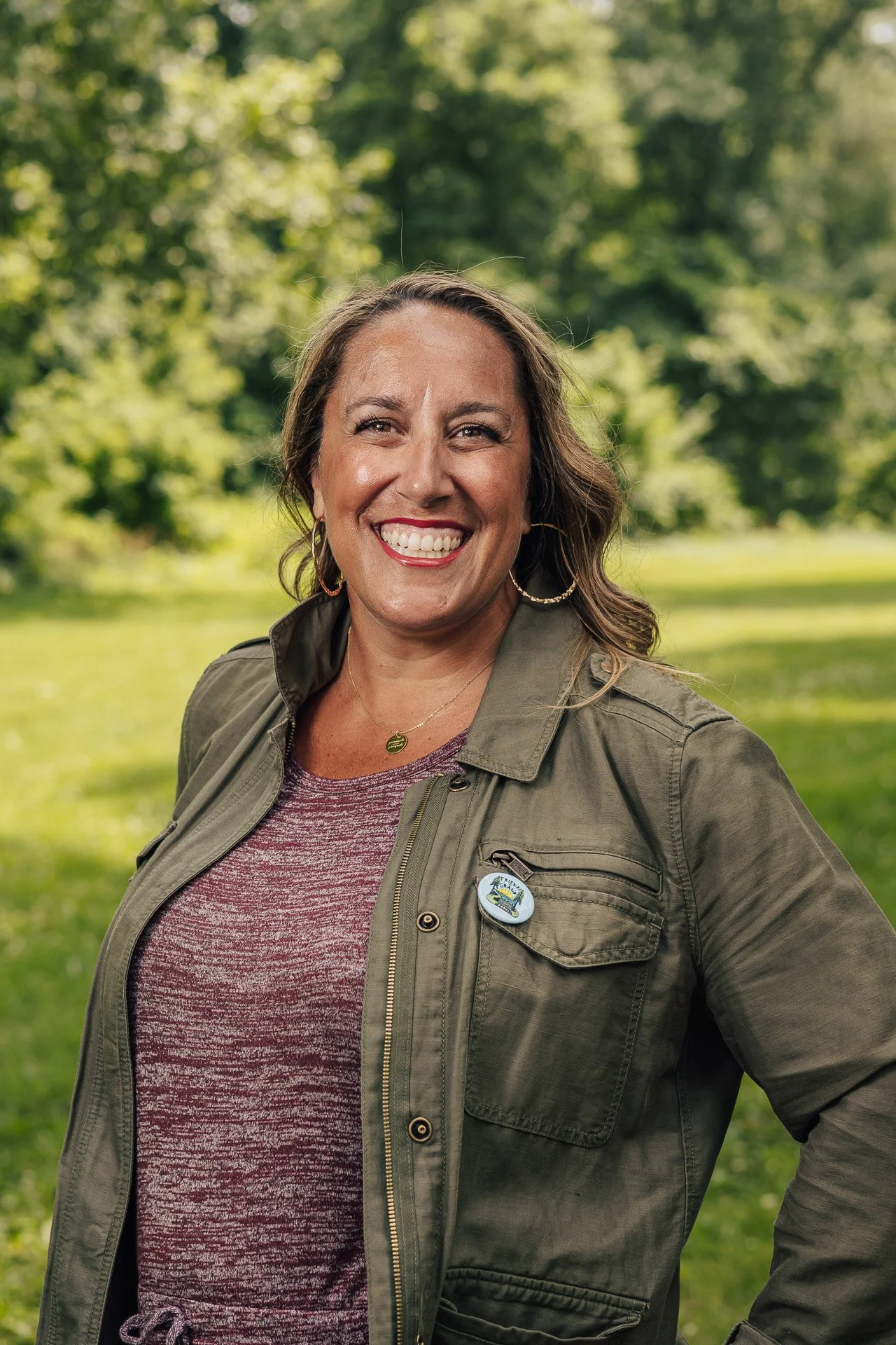 A woman smiling outdoors in a park, wearing a green jacket and maroon top, with a badge on her jacket.