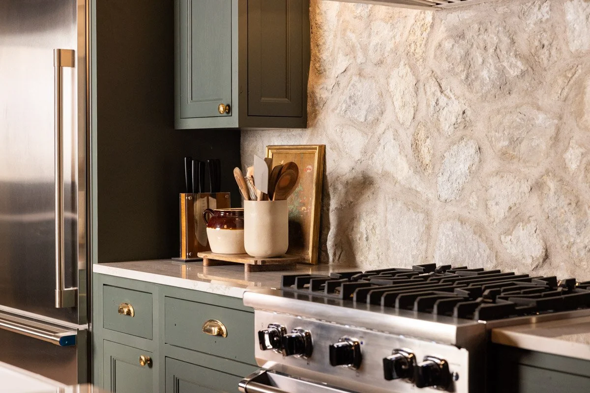 Kitchen with green cabinets, a stone backsplash, a stainless steel refrigerator, and a stove with black knobs. There are containers with cooking utensils and a framed picture on the counter.