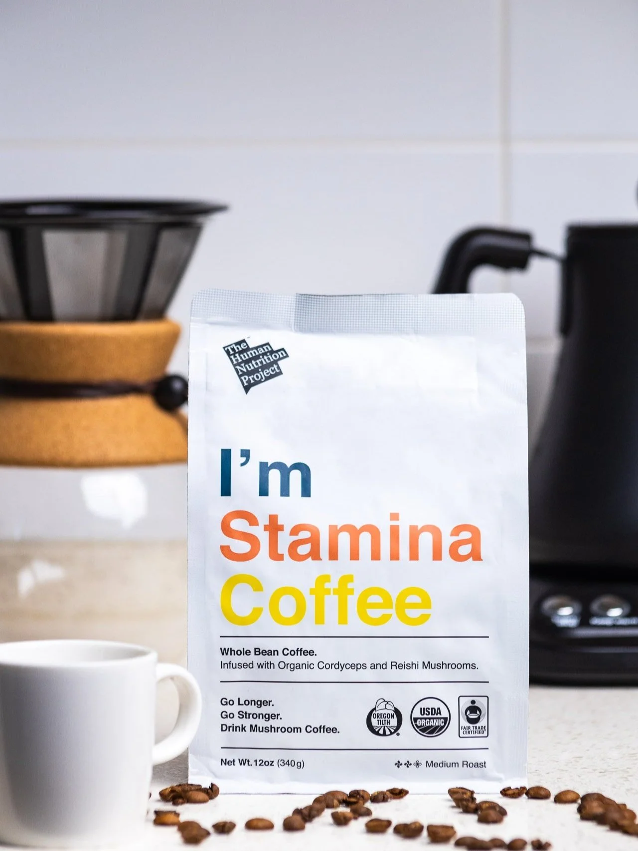 A package of whole bean mushroom coffee labeled 'I'm Stamina Coffee' sits on a counter with scattered coffee beans. A white coffee mug is partially visible in front of the package. In the background, kitchen appliances and a white tiled wall are visi