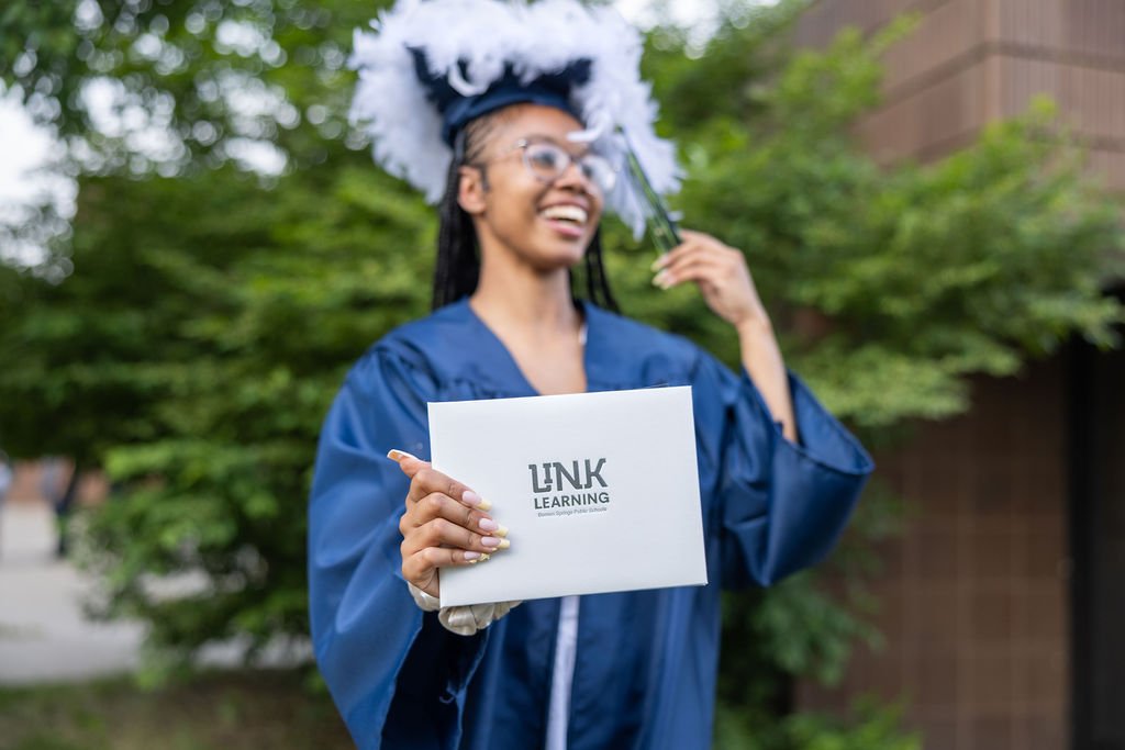 A woman in a blue graduation gown and cap, holding a diploma with 'LINK LEARNING' printed on it, standing outside with greenery in the background, smiling.