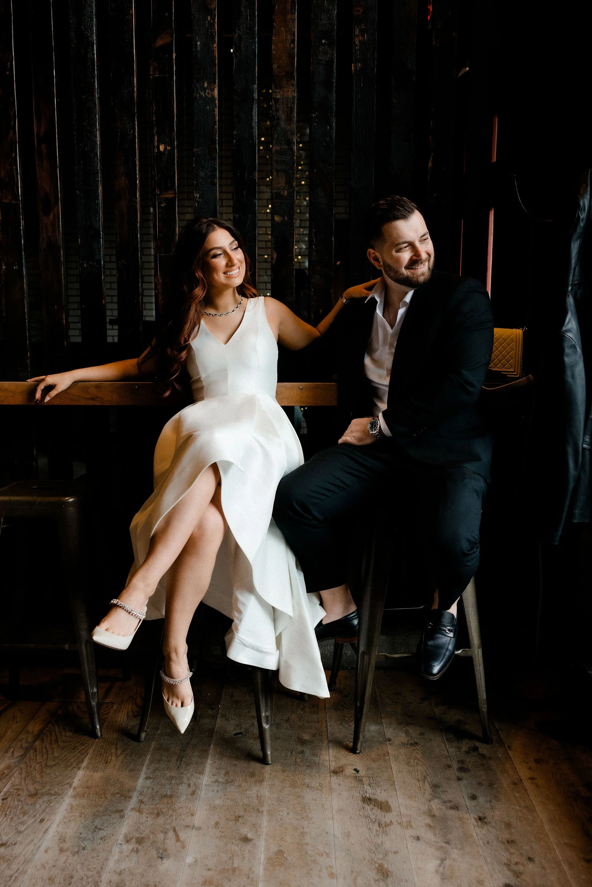A couple, dressed formally, sitting on chairs and smiling, with a dark wooden wall in the background.