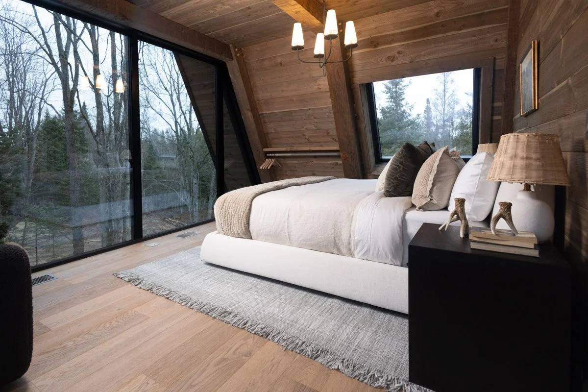 A modern bedroom with wooden walls and ceiling, a large glass window overlooking trees, a white bed with pillows, a black nightstand with a lamp, and a light fixture hanging from the ceiling.