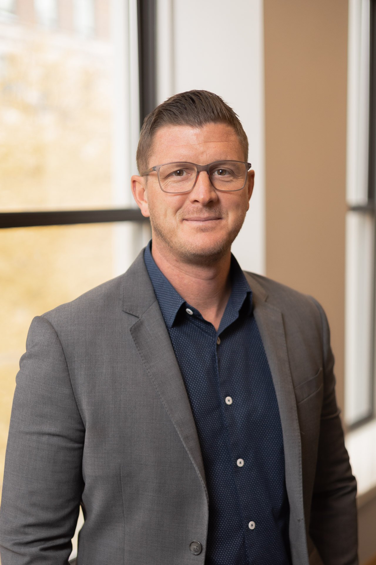 A man wearing glasses, a gray suit jacket, and a navy blue shirt standing indoors near windows with blurred autumn trees outside.