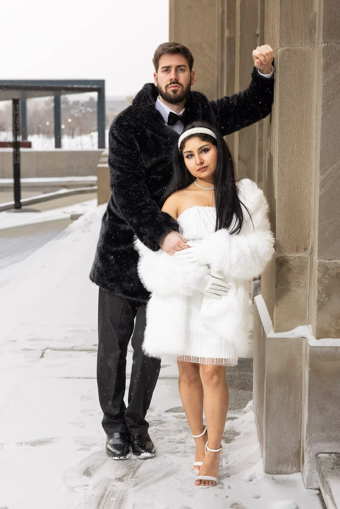 A man and woman in formal attire standing outside on a snowy day. The man is wearing a black tuxedo with a white shirt and black bow tie. The woman is dressed in a white off-the-shoulder dress, white gloves, and a white fur coat, with a pearl headban