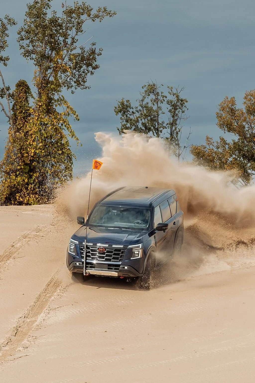 A black SUV driving through sandy terrain, kicking up dust with trees in the background and a cloudy sky.
