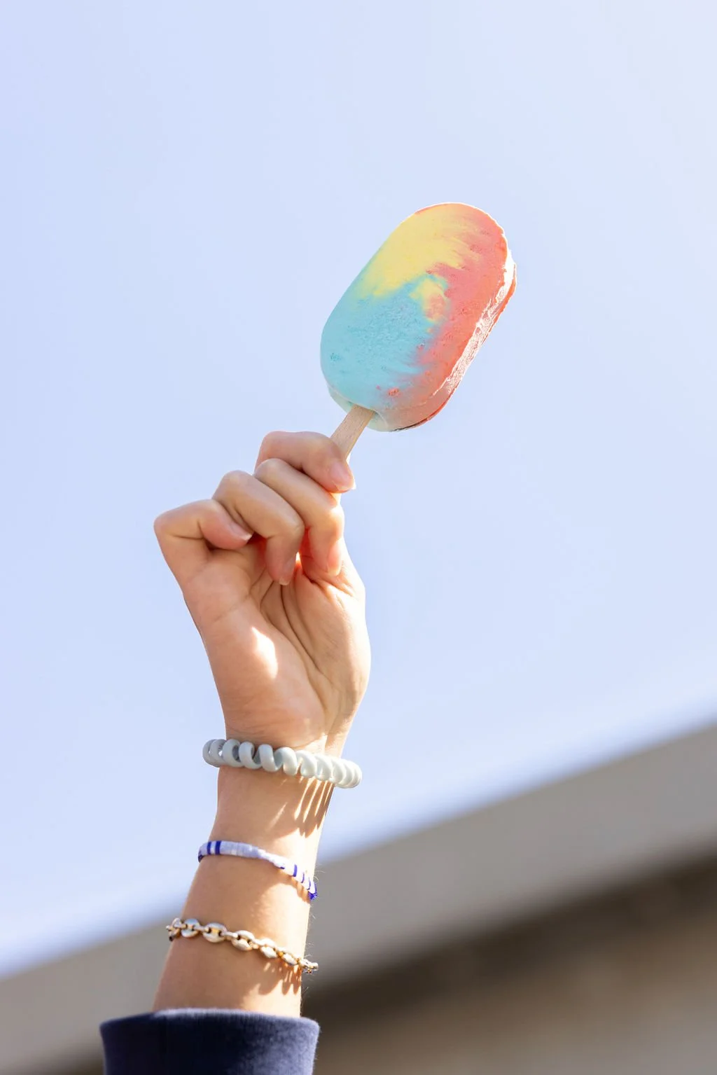 Hand holding a colorful rainbow pastel ice cream bar against a clear blue sky.