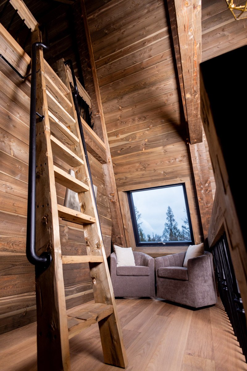A cozy corner in a wooden cabin with a wooden ladder leading up to a loft, a large window showing trees outside, and two beige armchairs with pillows arranged in an L-shape.