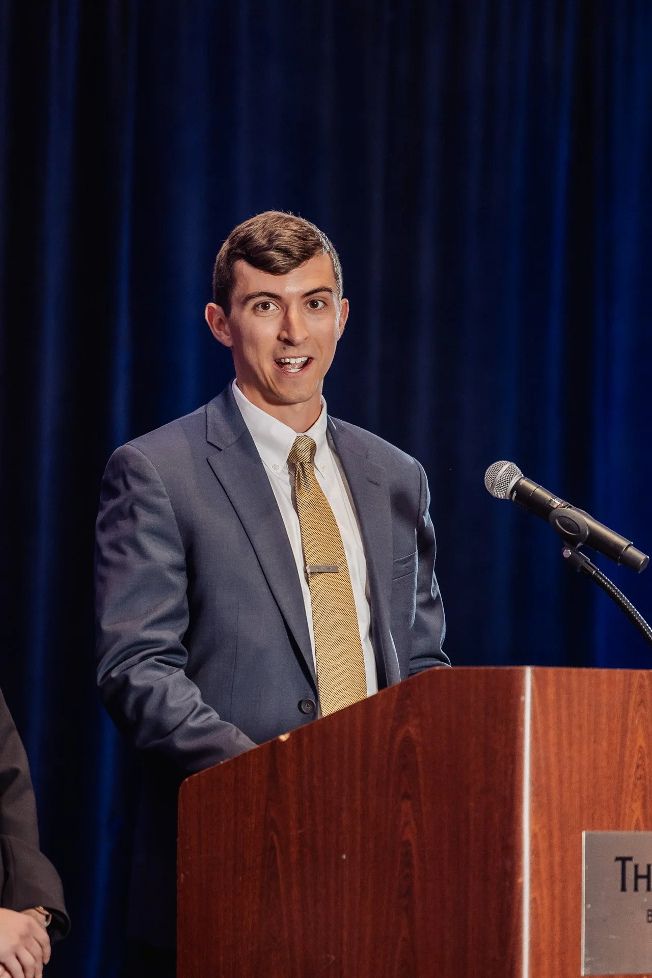 A young man in a gray suit, white shirt, and gold tie speaking at a wooden podium with a microphone, with a dark blue curtain background.