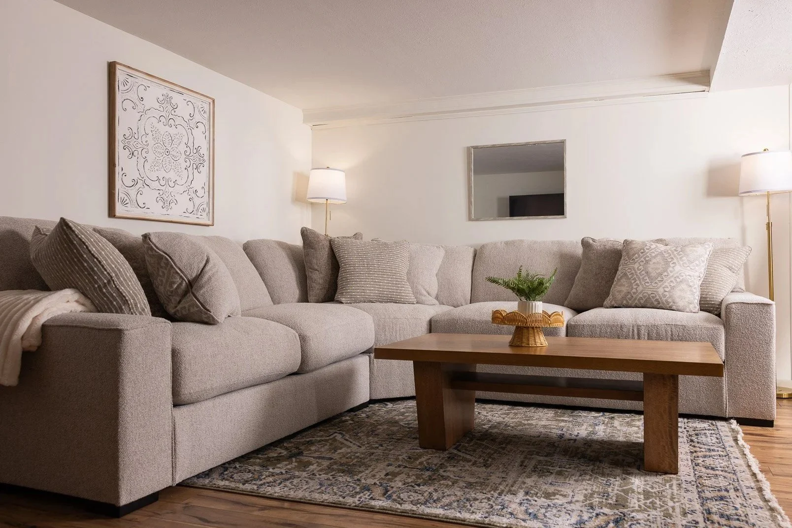 Living room with beige sectional sofa, decorative pillows, a wooden coffee table with a potted plant, two floor lamps, a wall picture, and a mirror, on a patterned rug.