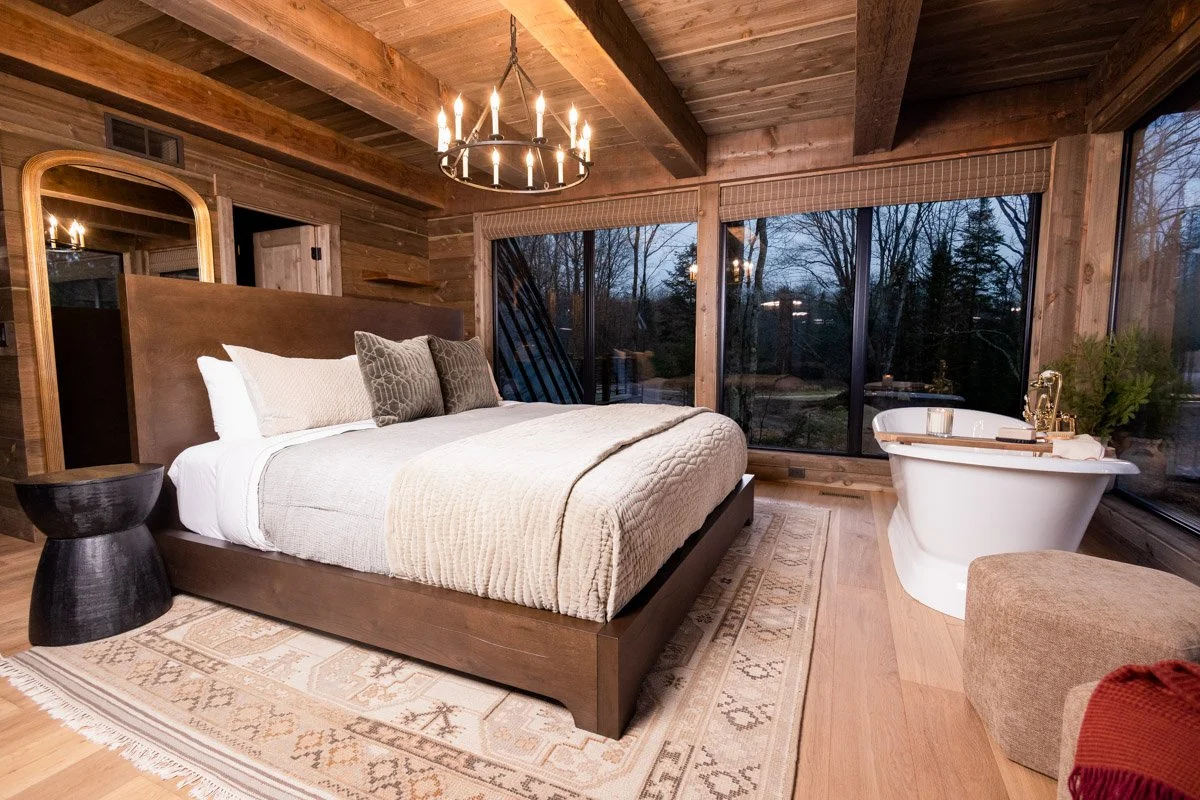 A cozy bedroom with wooden walls and ceiling, a large bed with neutral-colored bedding and throw pillows, a circular chandelier, large windows with a view of trees outside, and a freestanding bathtub with a gold faucet near the window.
