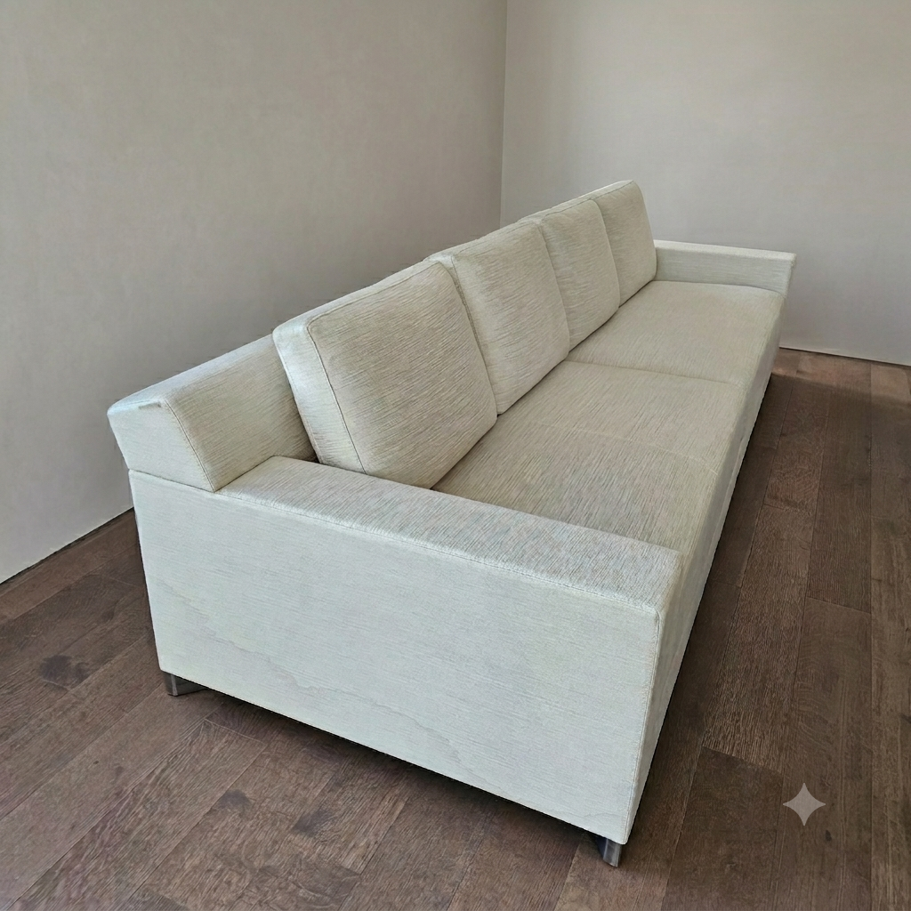 A light beige modern sofa with three cushions on a dark hardwood floor against a plain white wall.