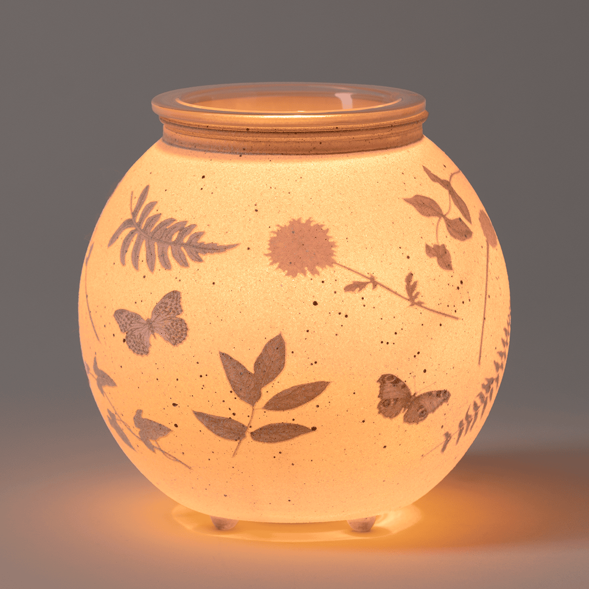A soft, ambient glow highlights the sanded glass and delicate botanical and butterfly details of the Veiled Meadow Warmer.