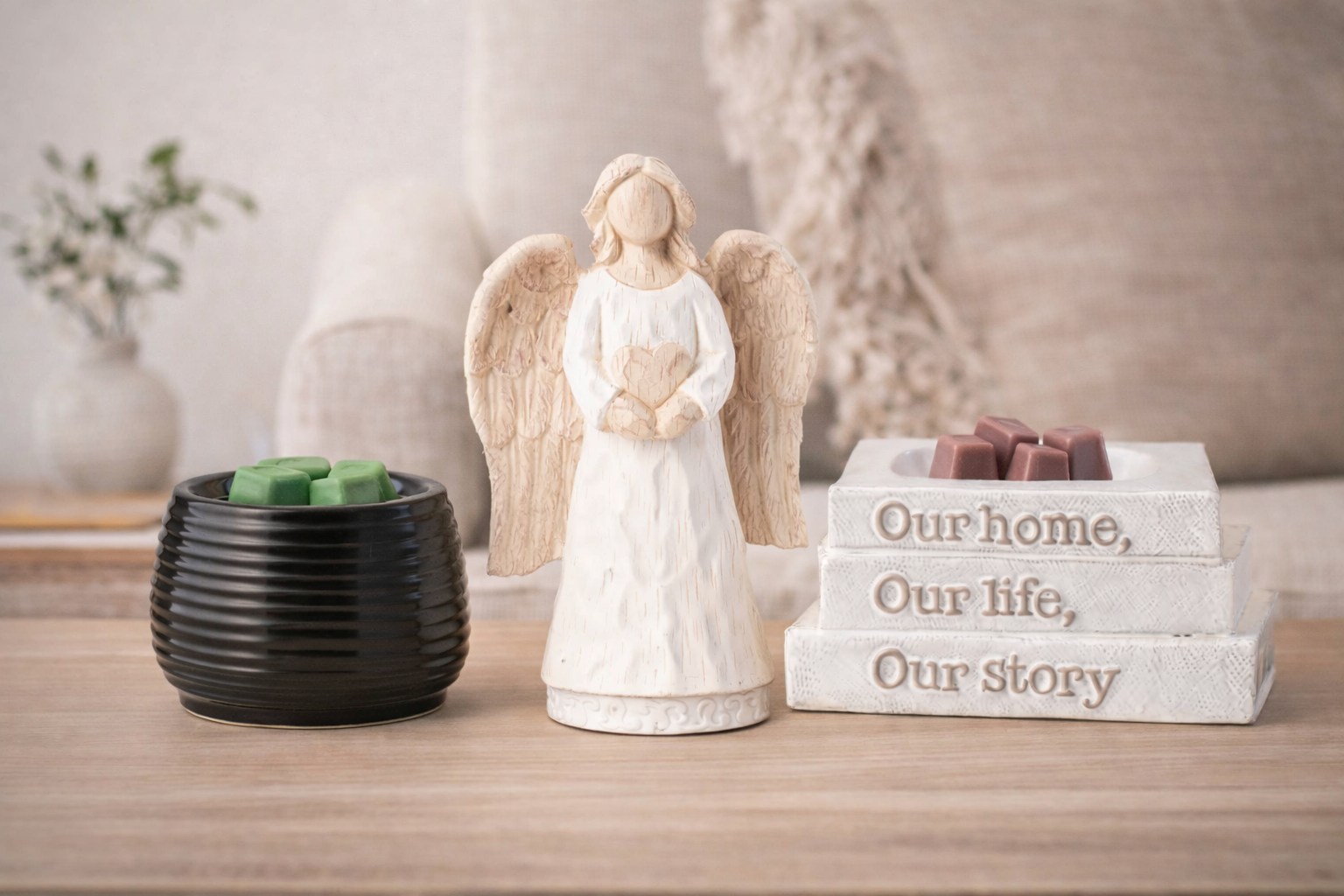 Three Scentsy element wax warmers displayed on a wooden surface in a modern cozy home setting, including a black ceramic warmer, an angel figurine warmer, and a white book-style warmer with wax cubes