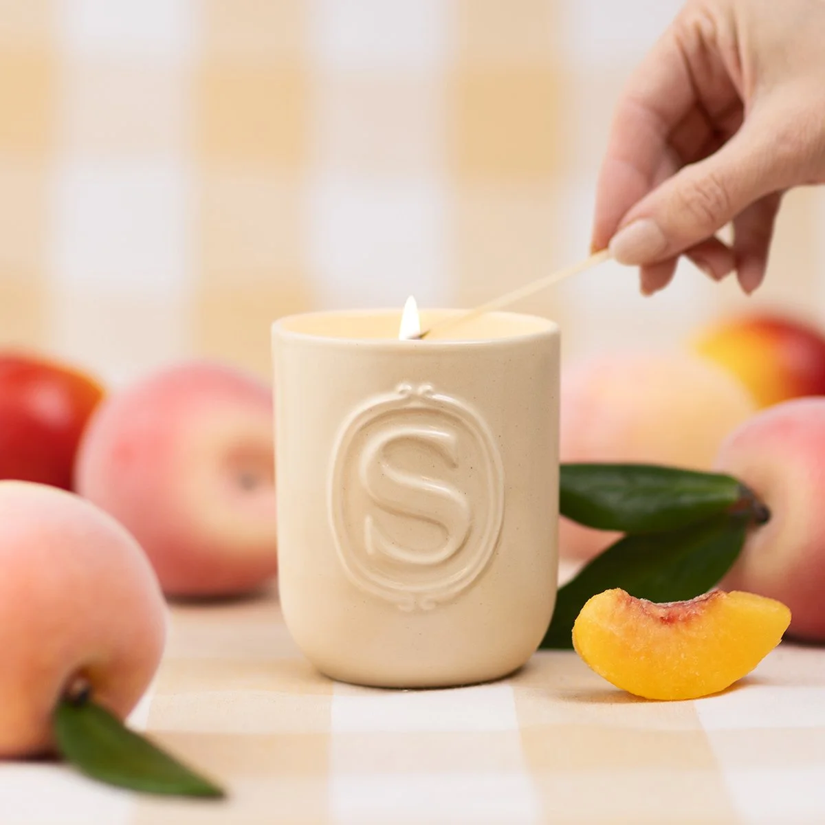 A Sunlit Peach Scentsy candle being lit beside fresh peaches, highlighting warm, sweet fruit-inspired fragrance in a soft kitchen setting.