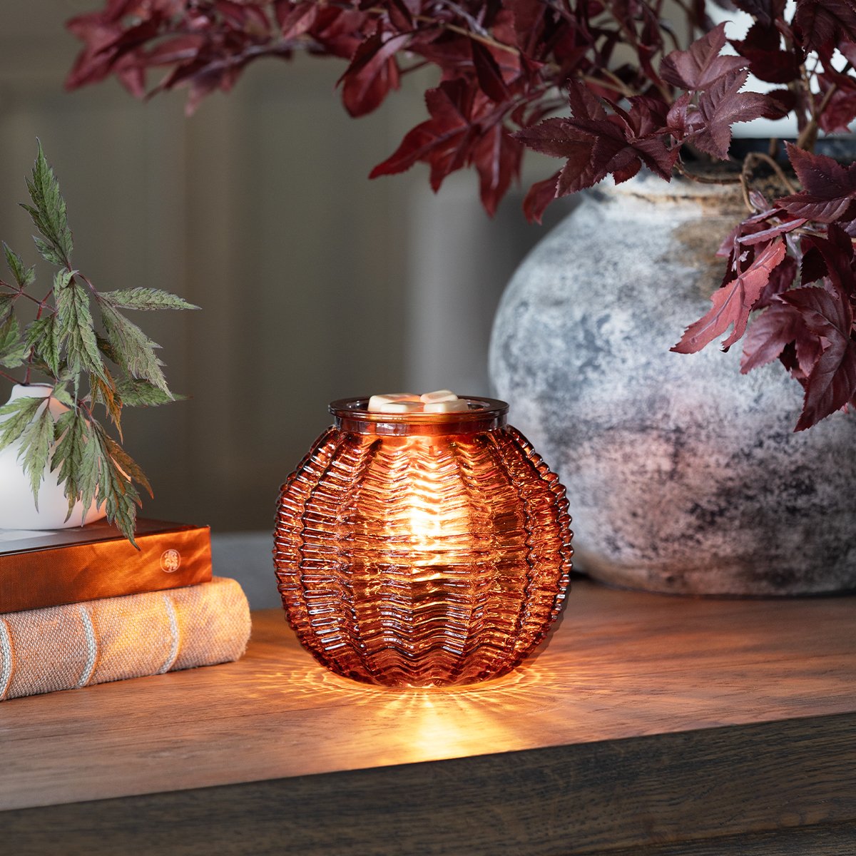 An amber glass Scentsy warmer with a sculptural ribbed design casts a warm, ambient glow on a wood surface styled with natural décor.